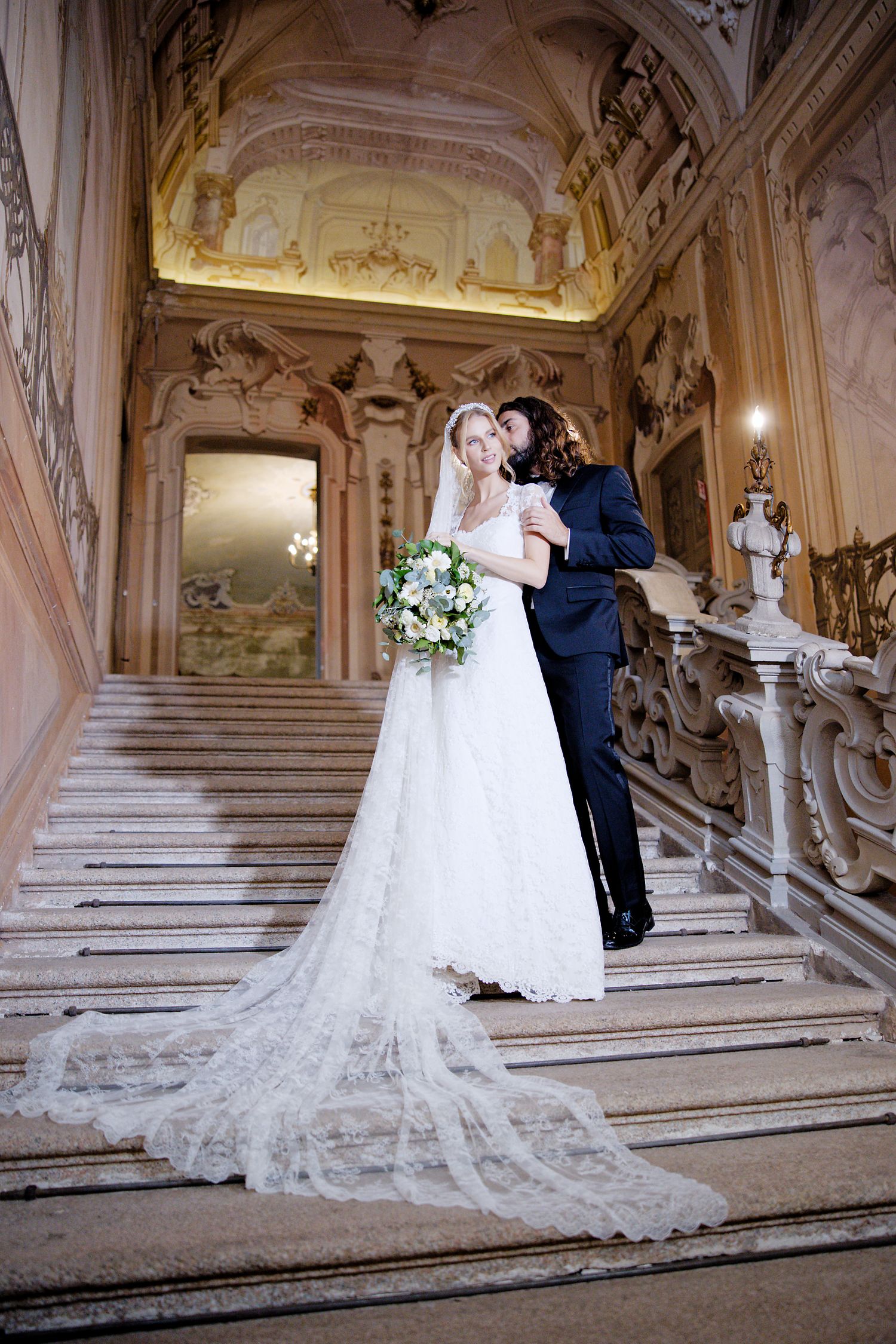 A wedding couple embraces on an ornate marble staircase in a luxurious palace with baroque architectural details.