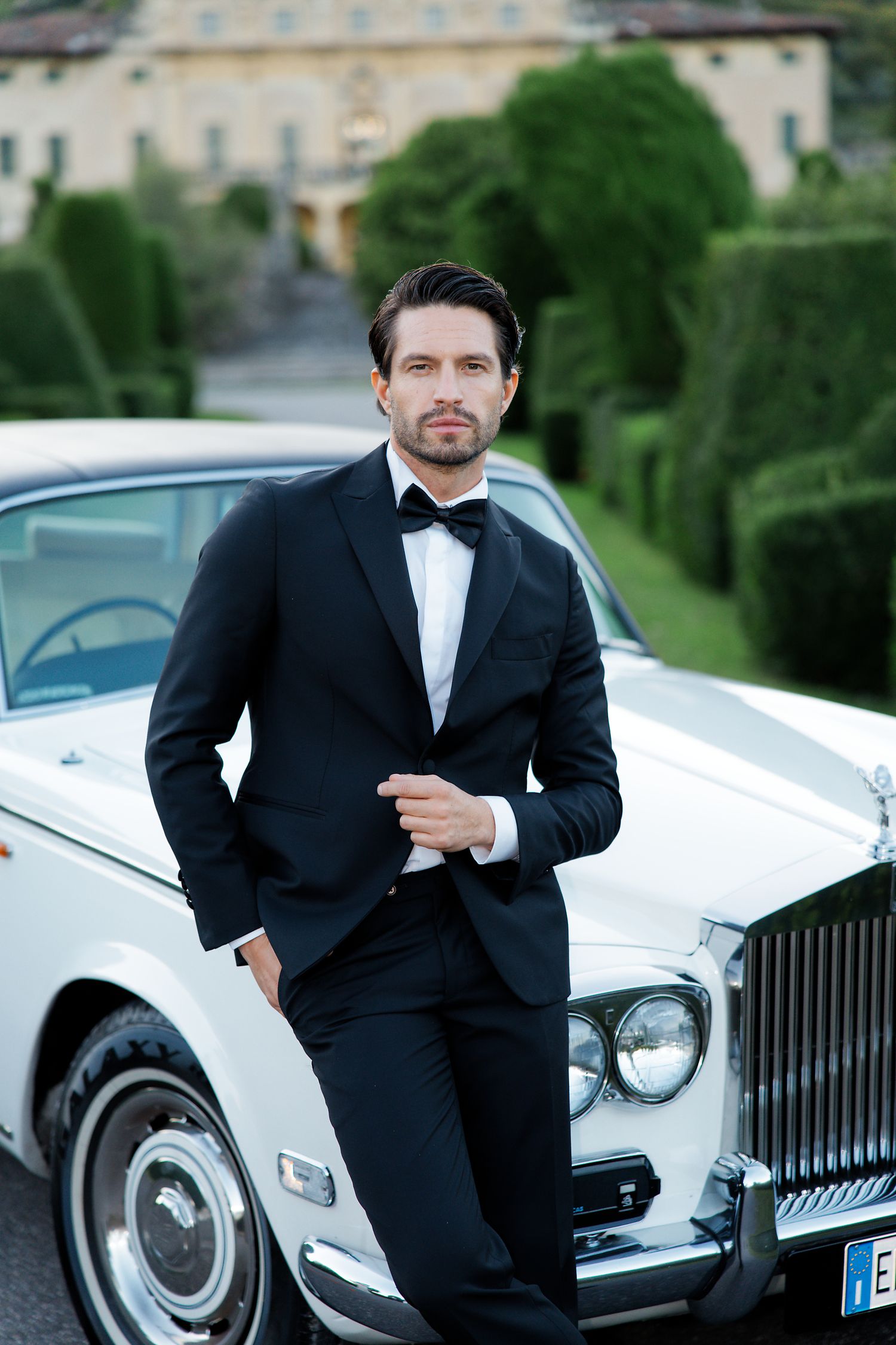 A well-dressed gentleman in a black tuxedo poses next to a classic white Rolls-Royce car in an elegant garden setting.