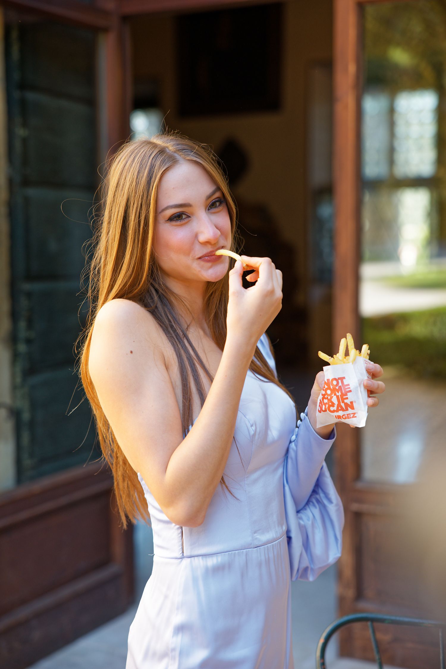 Woman in white dress eating french fries while standing near a window.