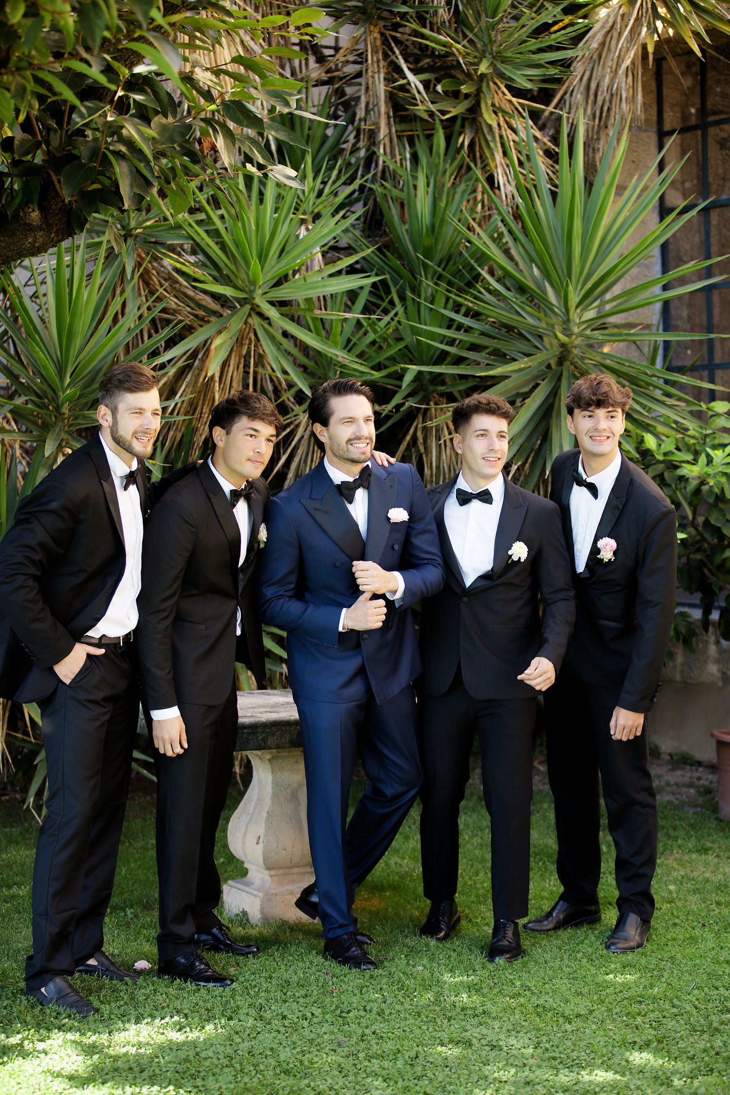 Groomsmen in formal tuxedos pose together against tropical plants in garden setting.