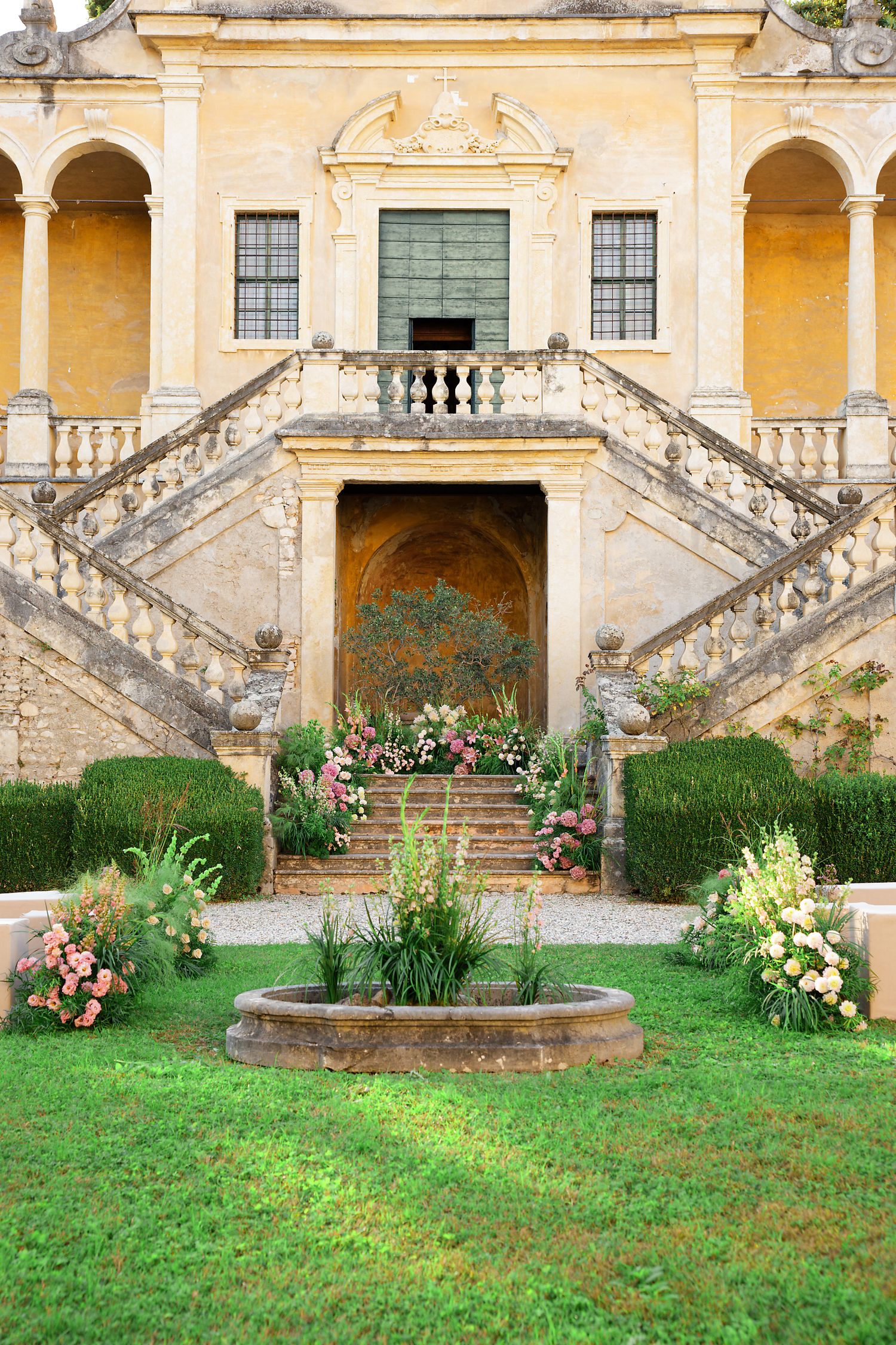 Elegant villa entrance with symmetrical double staircase, yellow walls and ornate architectural details in a manicured garden setting.