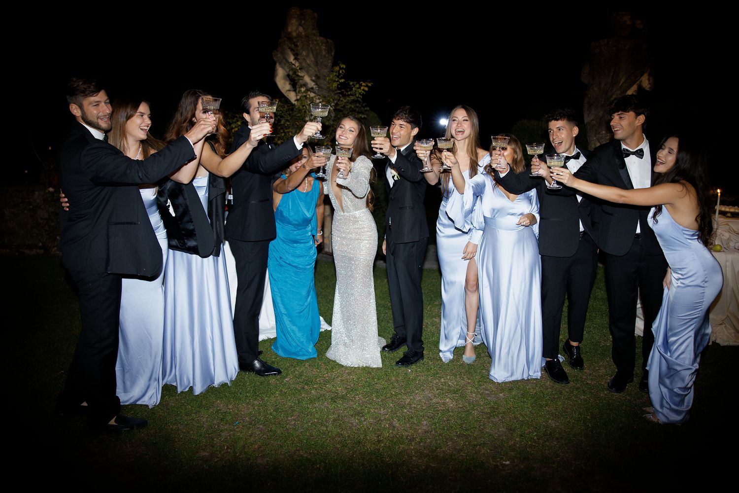 Wedding party guests in formal attire gather for a celebratory nighttime photo on a lawn.