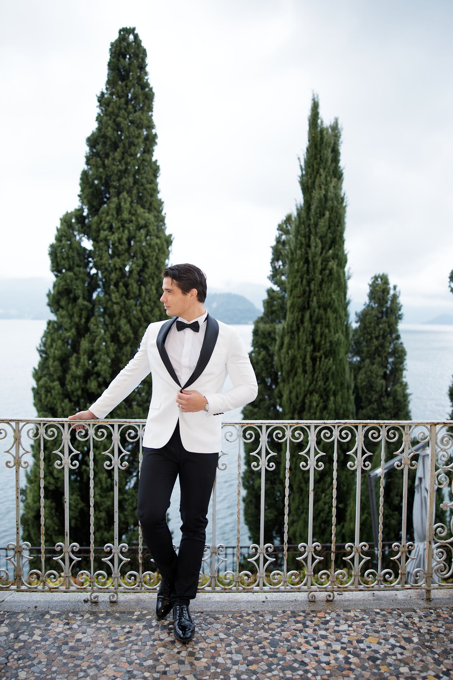 Groom in white tuxedo jacket poses against iron railing with lake and cypress trees.