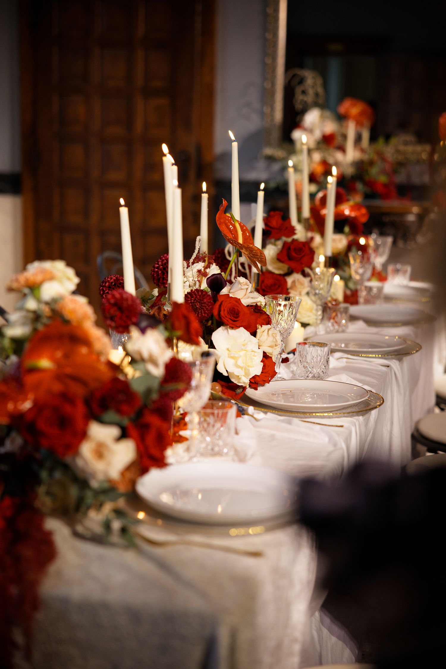 Elegant wedding table decorated with red and white flowers, tall candles, and gold-rimmed plates.