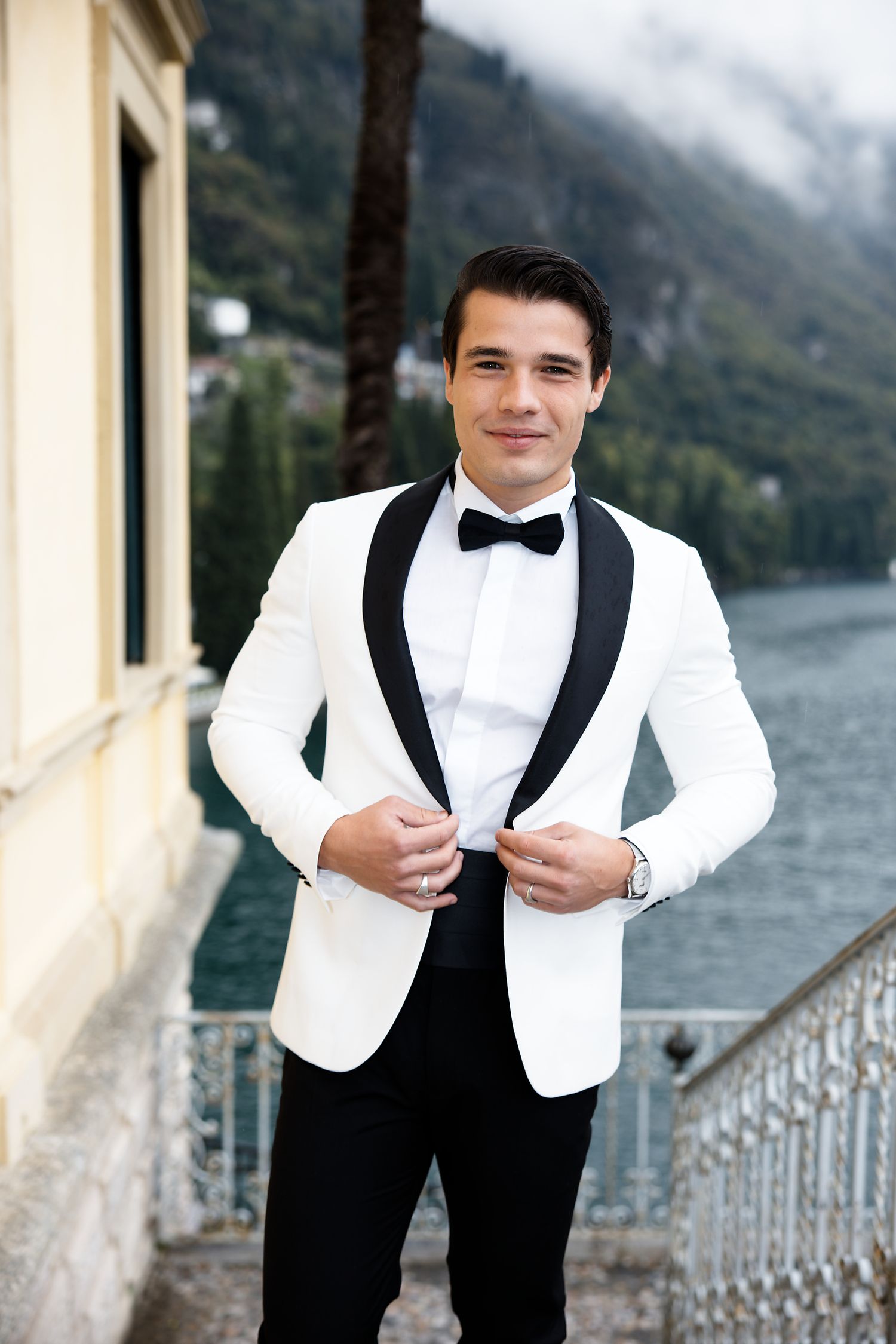 Elegant groom in white dinner jacket with black bow tie poses against scenic lake and mountain backdrop.