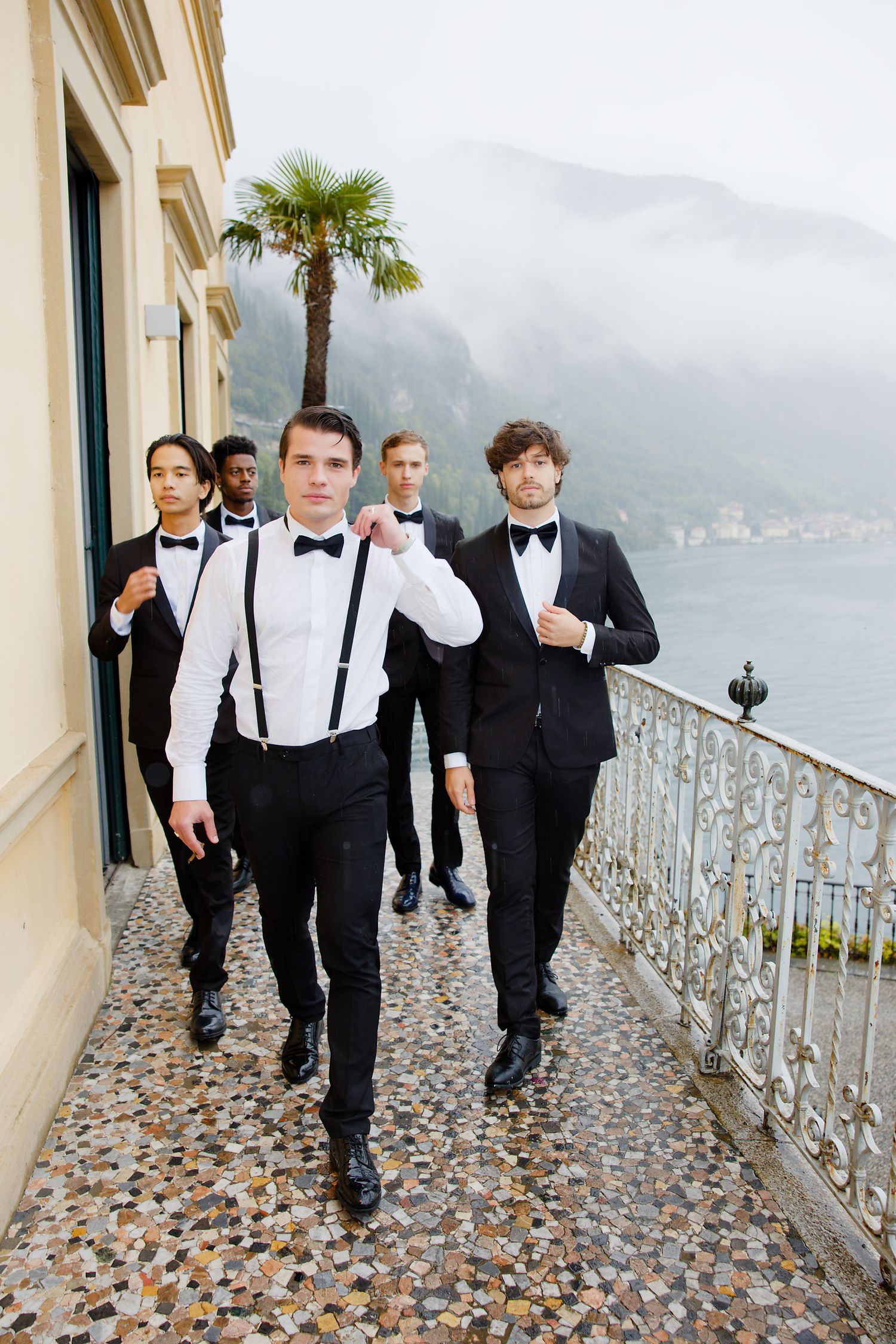 Wedding party in formal attire walking along scenic coastal balcony with palm trees.