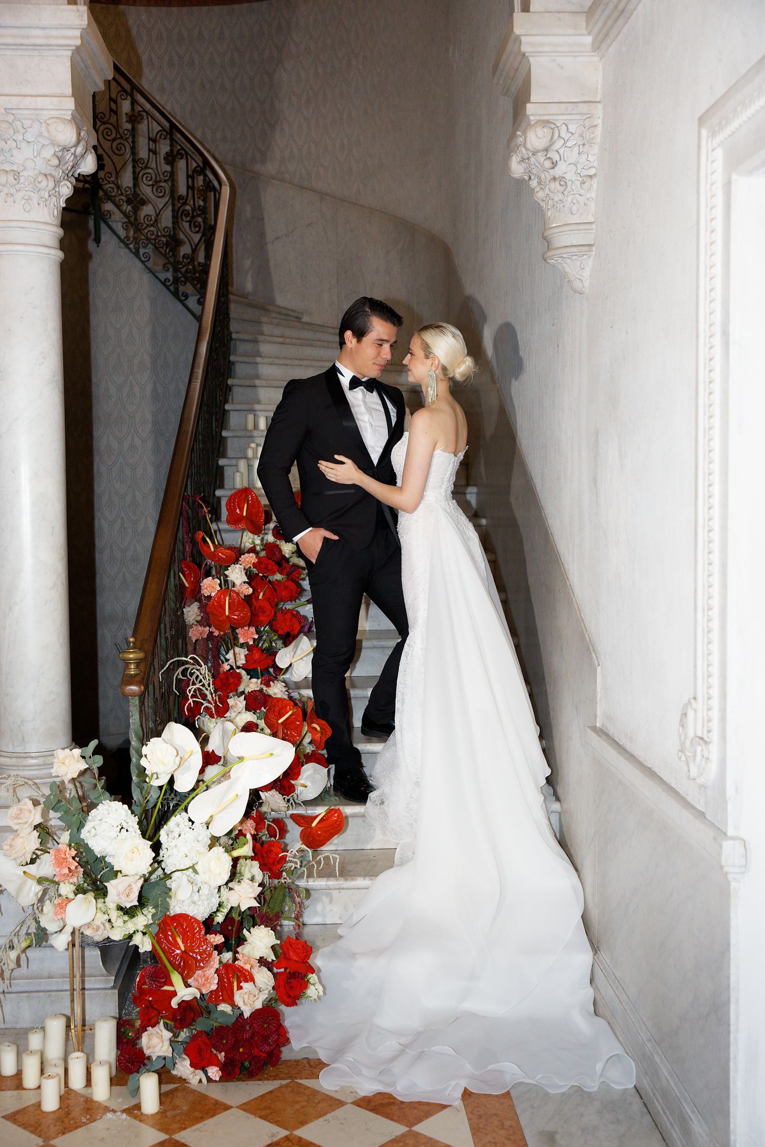Romantic wedding portrait on grand staircase decorated with red and white floral arrangements.