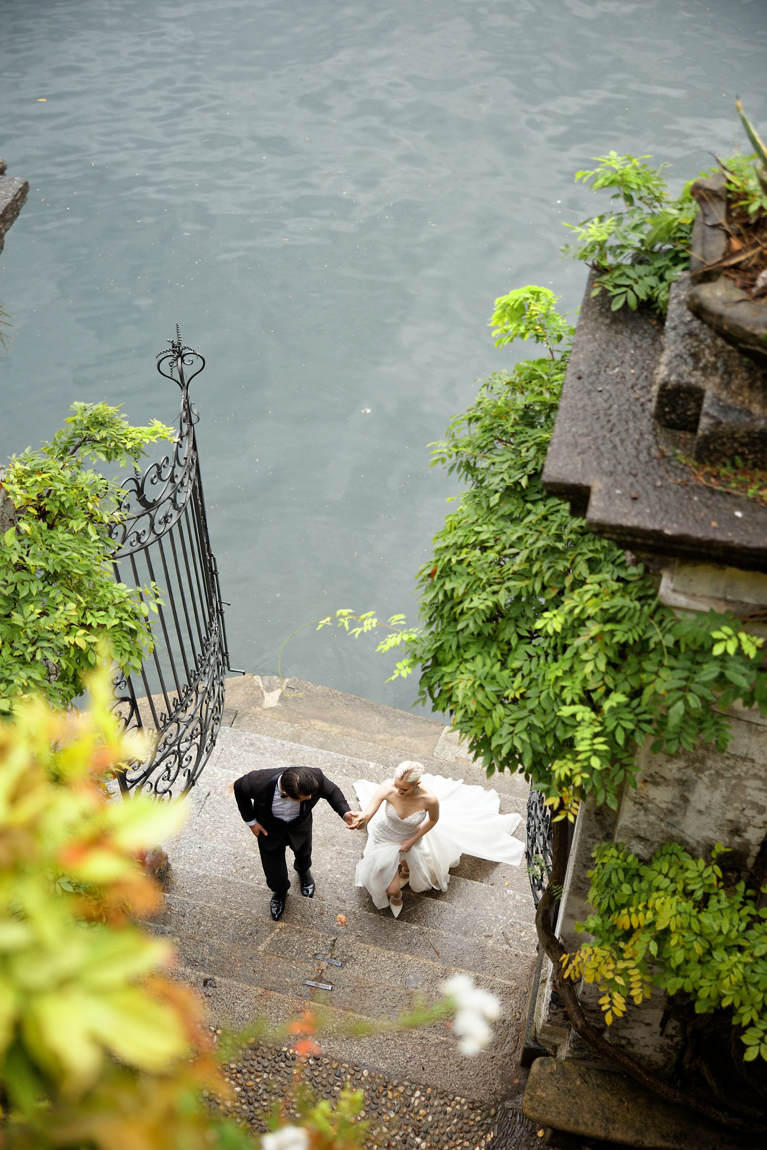 Aerial view of a couple walking down stone steps near water surrounded by lush greenery.