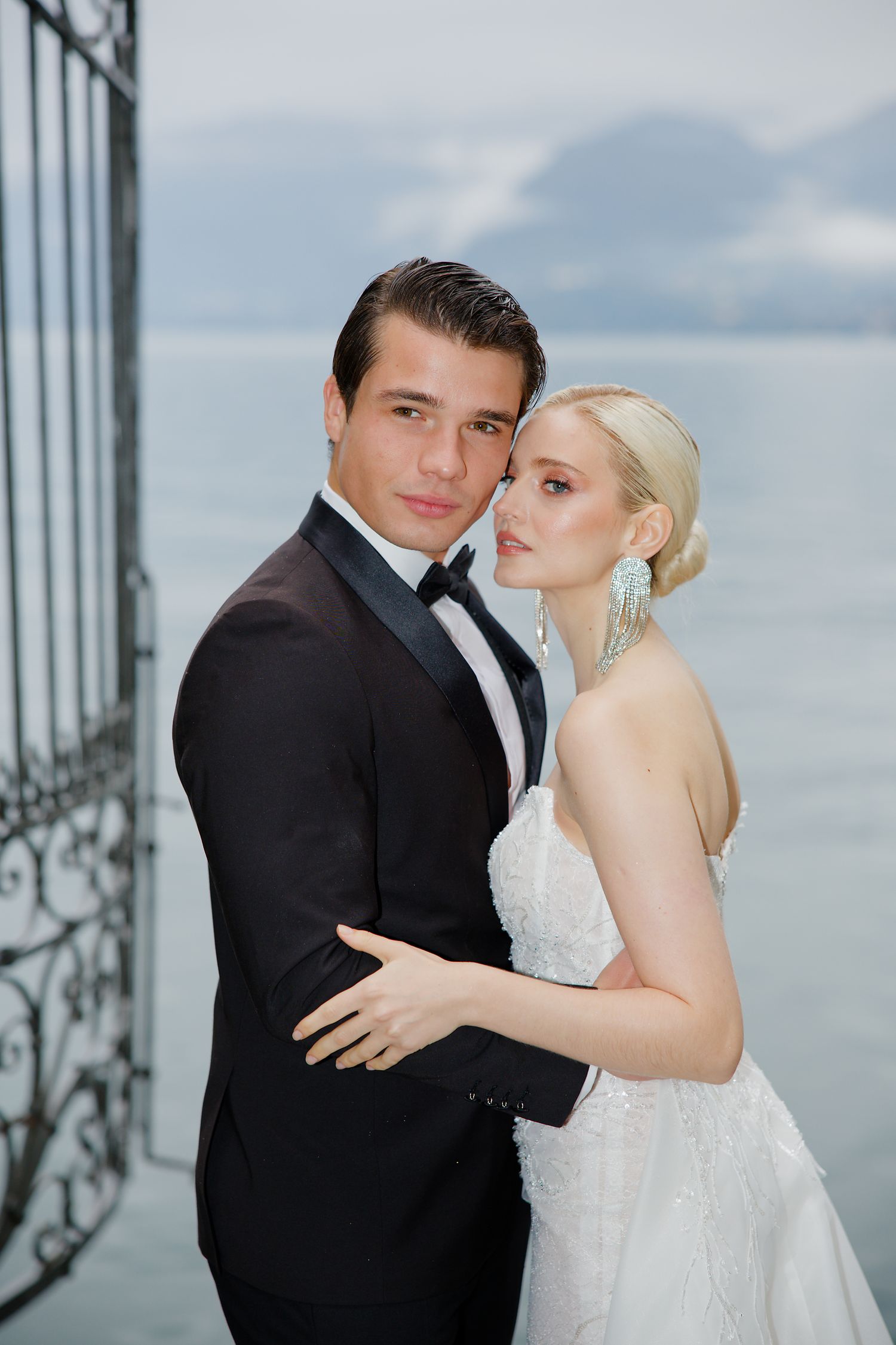 Elegant couple in formal wedding attire pose on balcony overlooking misty lake views.