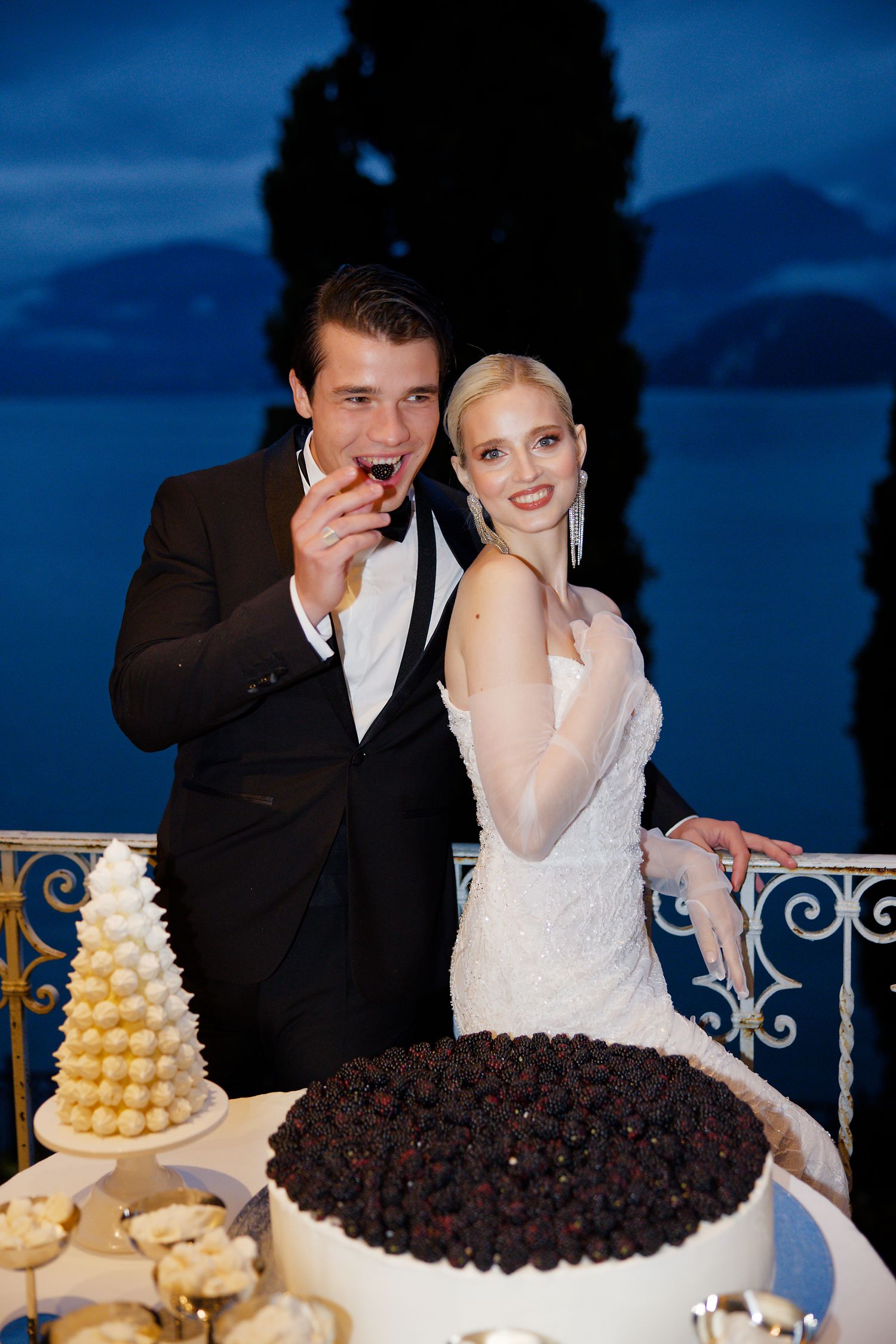 A bride and groom enjoy their wedding cake display featuring a tall yellow macaron tower and chocolate dessert on a balcony.