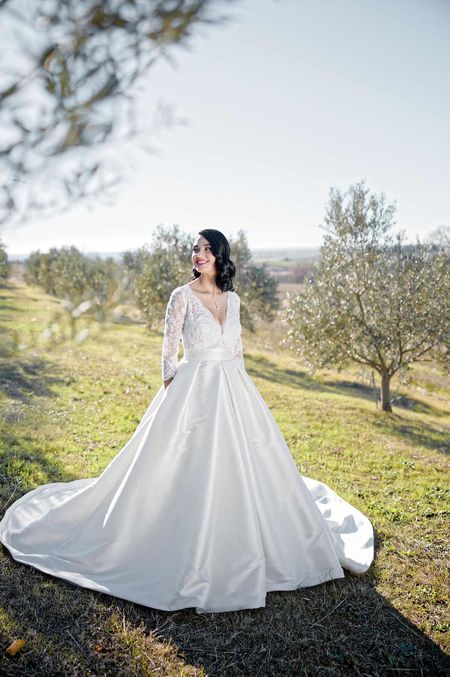 Elegant long-sleeved wedding gown with lace details and flowing train photographed in a sunlit olive grove.