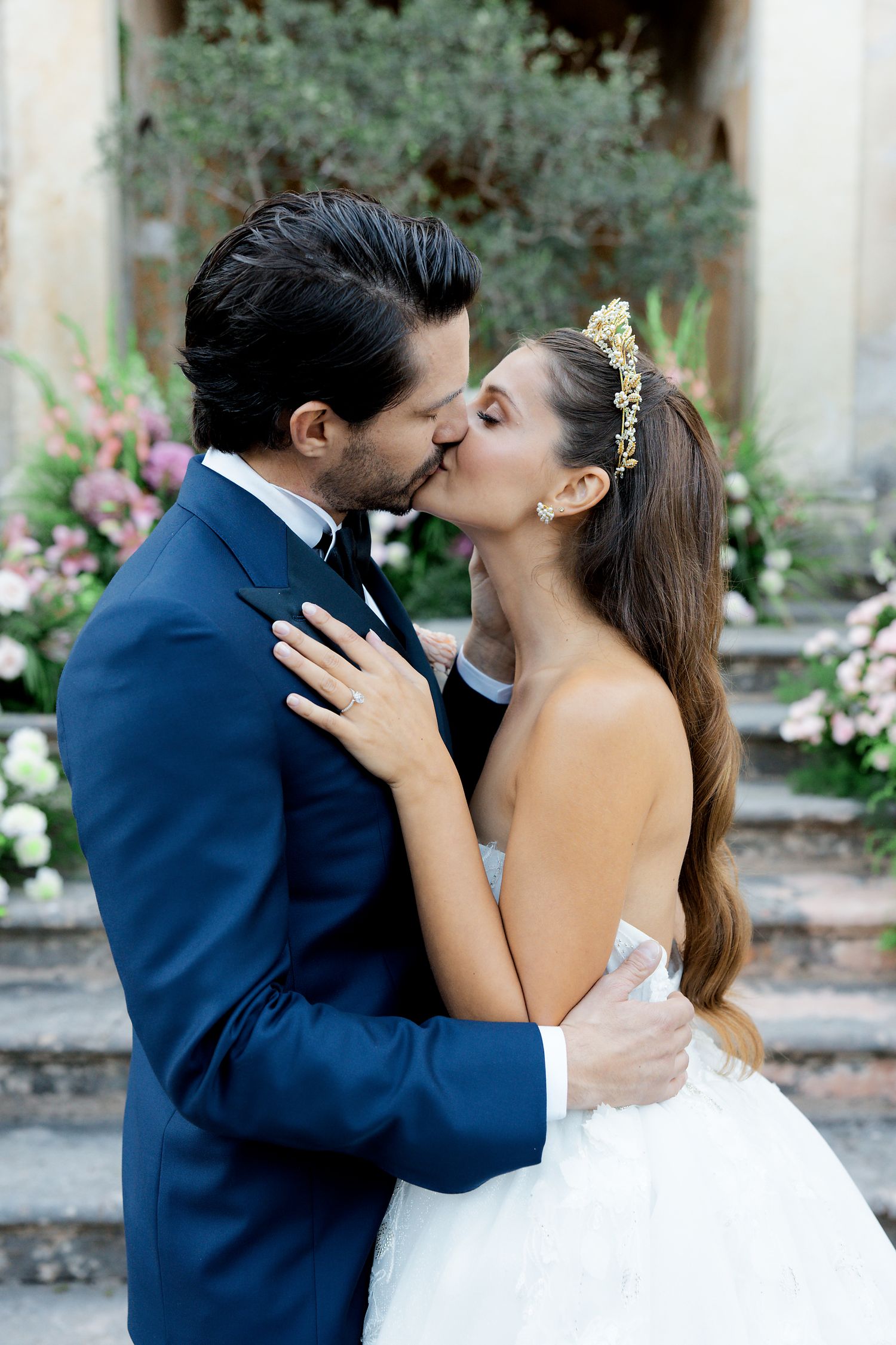 A romantic wedding couple shares a tender kiss while standing on stone steps with pink flowers in the background.
