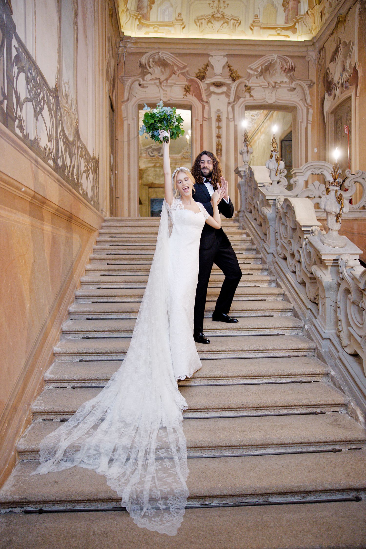 A bride in a lace wedding gown and groom in black suit pose playfully on an ornate grand staircase.