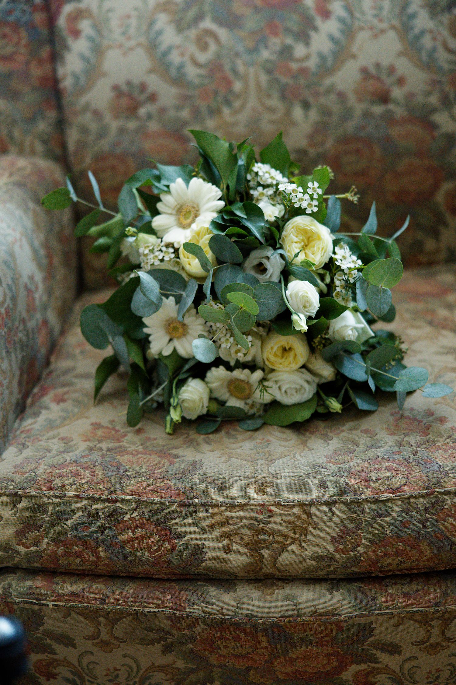 A delicate wedding bouquet with white roses, daisies and eucalyptus leaves rests on a floral patterned vintage chair.