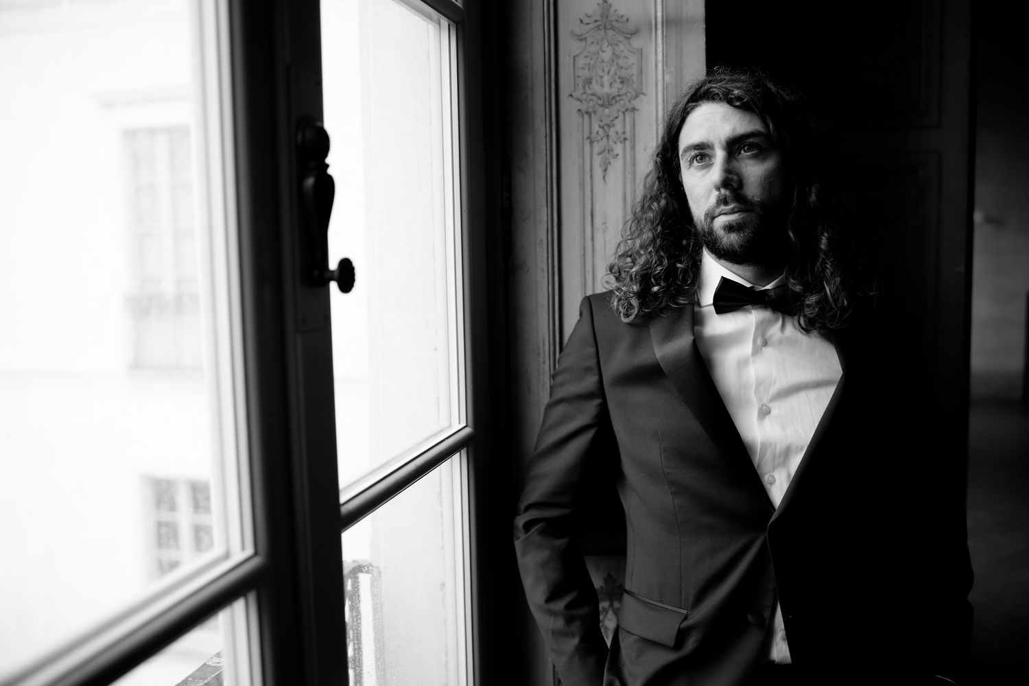 Black and white portrait of groom in tuxedo standing by window in dramatic lighting.
