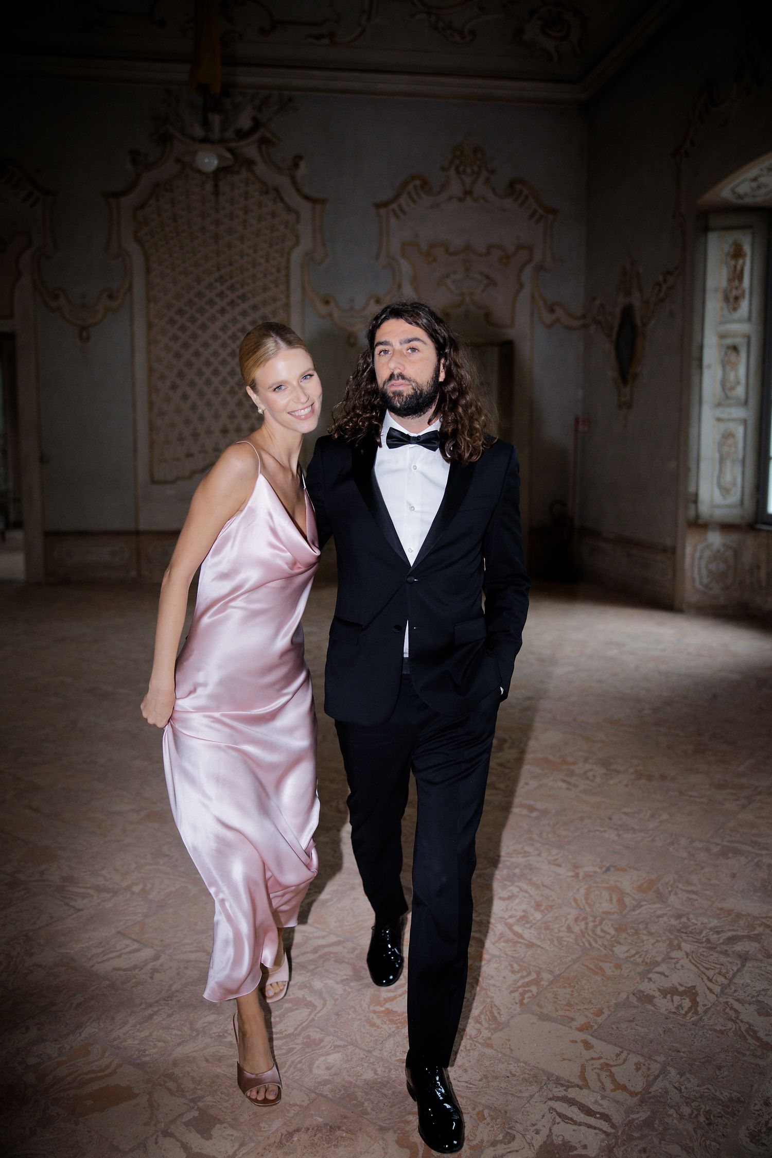 A pair walking together in formal attire through an elegant room with ornate wall detailing.