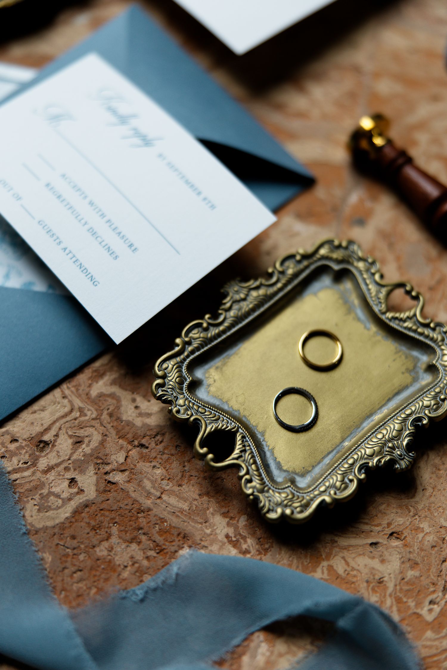 Wedding rings displayed on ornate gold tray with blue invitation and wax seal.