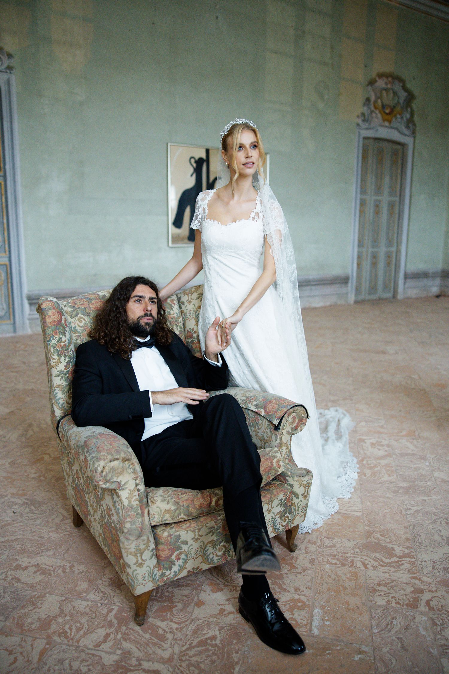 Bride in white lace gown and groom in black suit pose in vintage armchair at ornate venue.