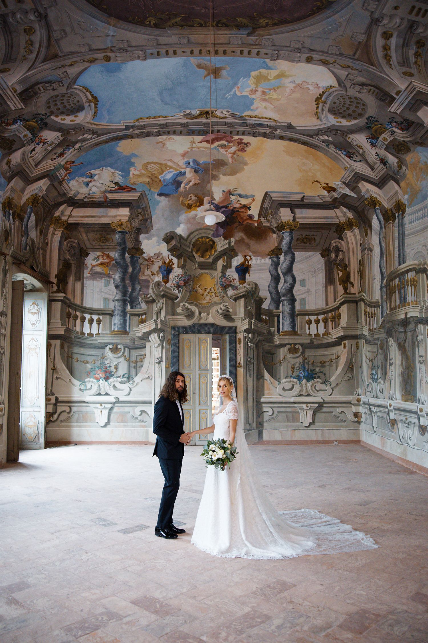 A couple stands in a grand baroque hall with ornate frescoed ceilings and classical architectural details.