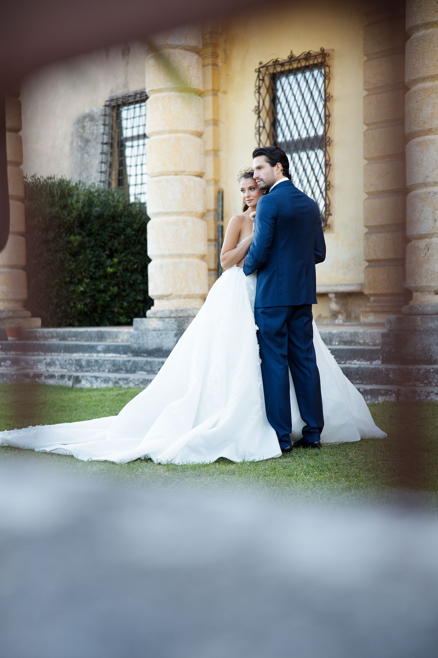 A romantic wedding portrait of a couple embracing on manicured grounds of a historic building.