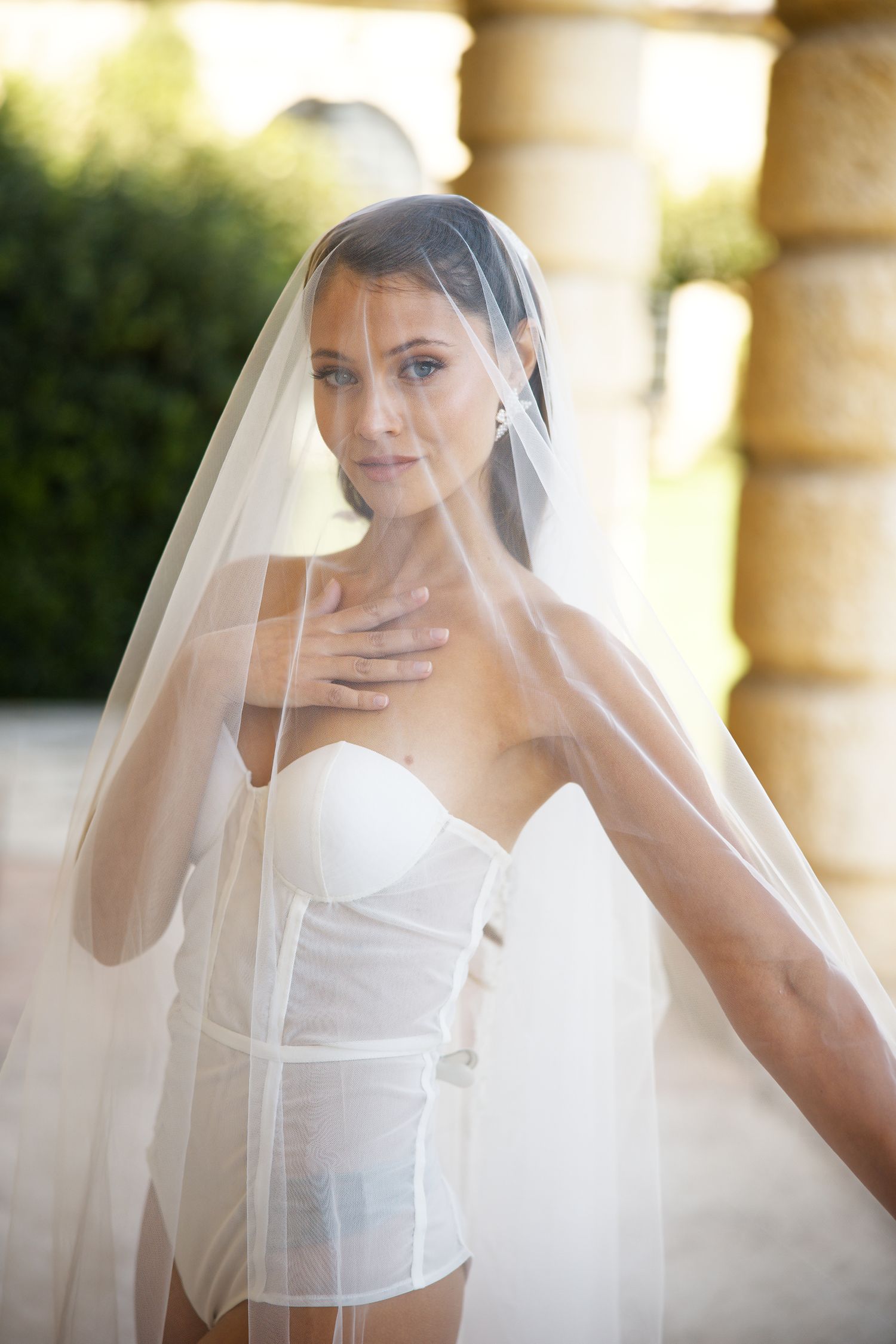 Bride in white corset and flowing veil poses elegantly in sunlit colonnade.