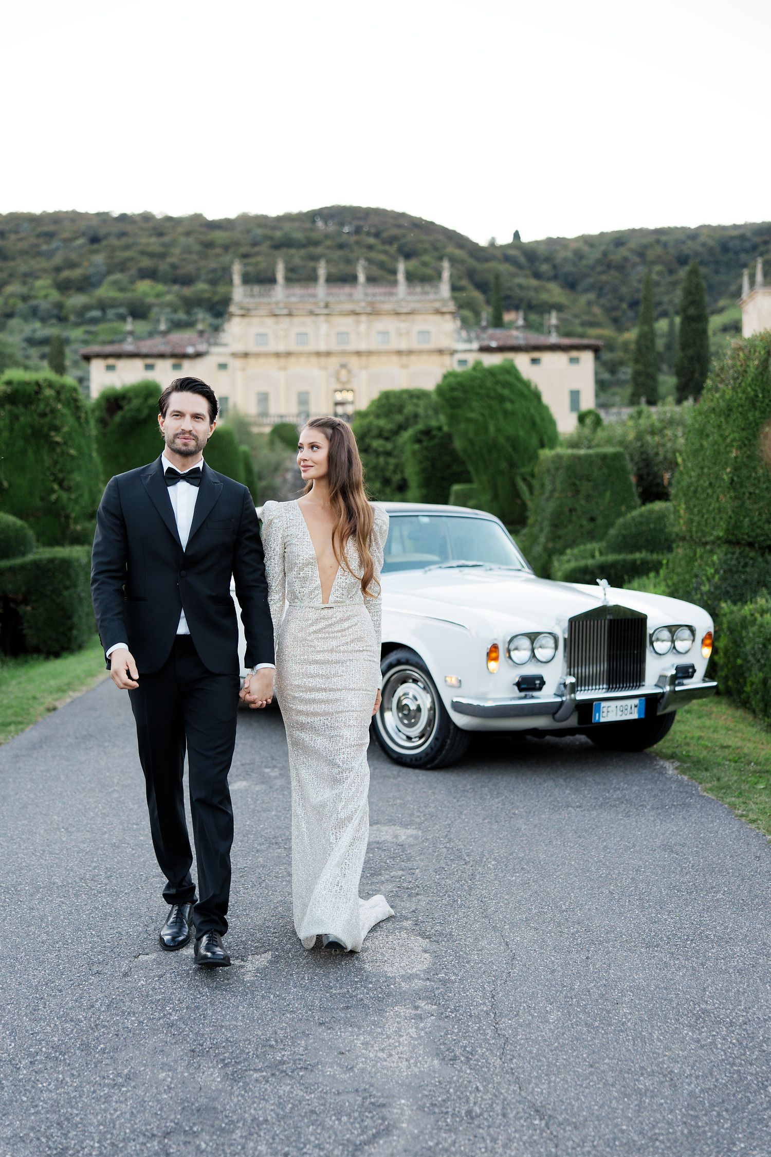 A vintage white Rolls Royce parked on a driveway of an elegant Italian villa with manicured gardens.