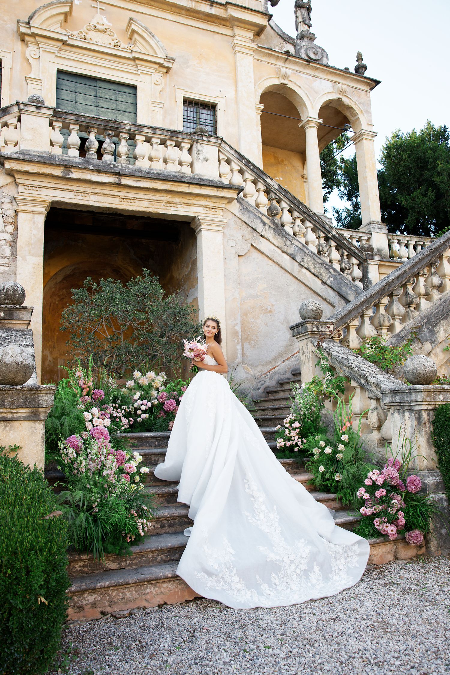 Elegant white wedding dress with long train photographed on historic stone staircase with blooming flowers and arches.
