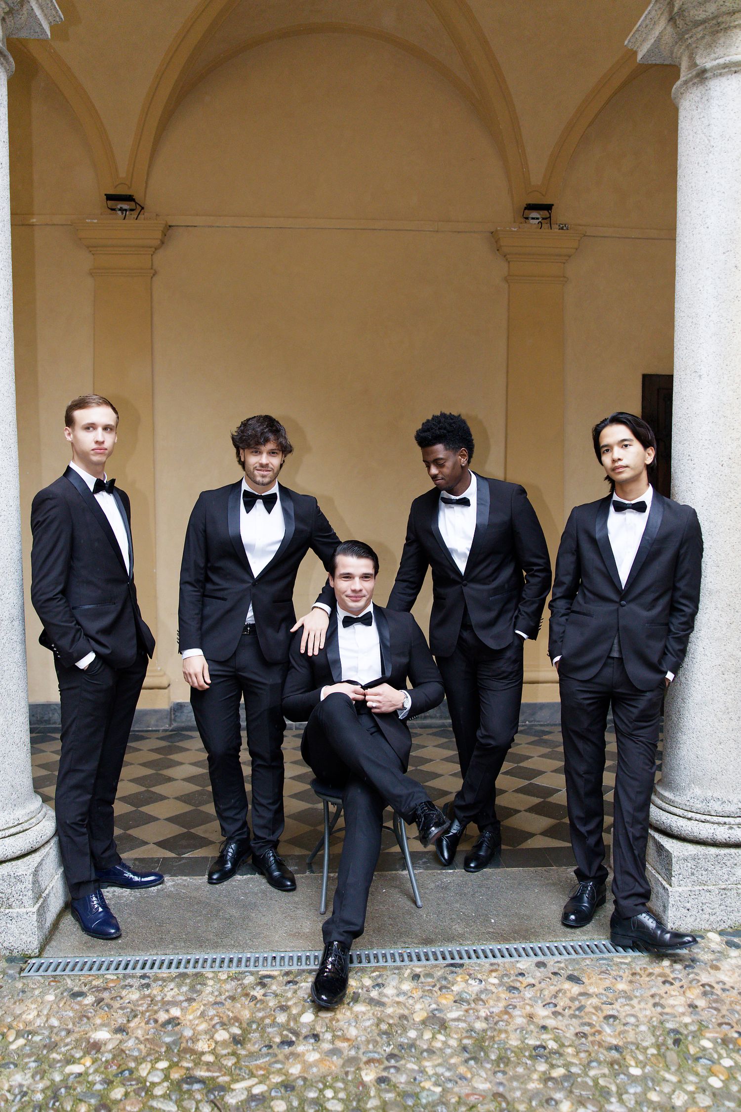 A group of formally dressed men in tuxedos pose elegantly under an arched colonnade with classic architecture.