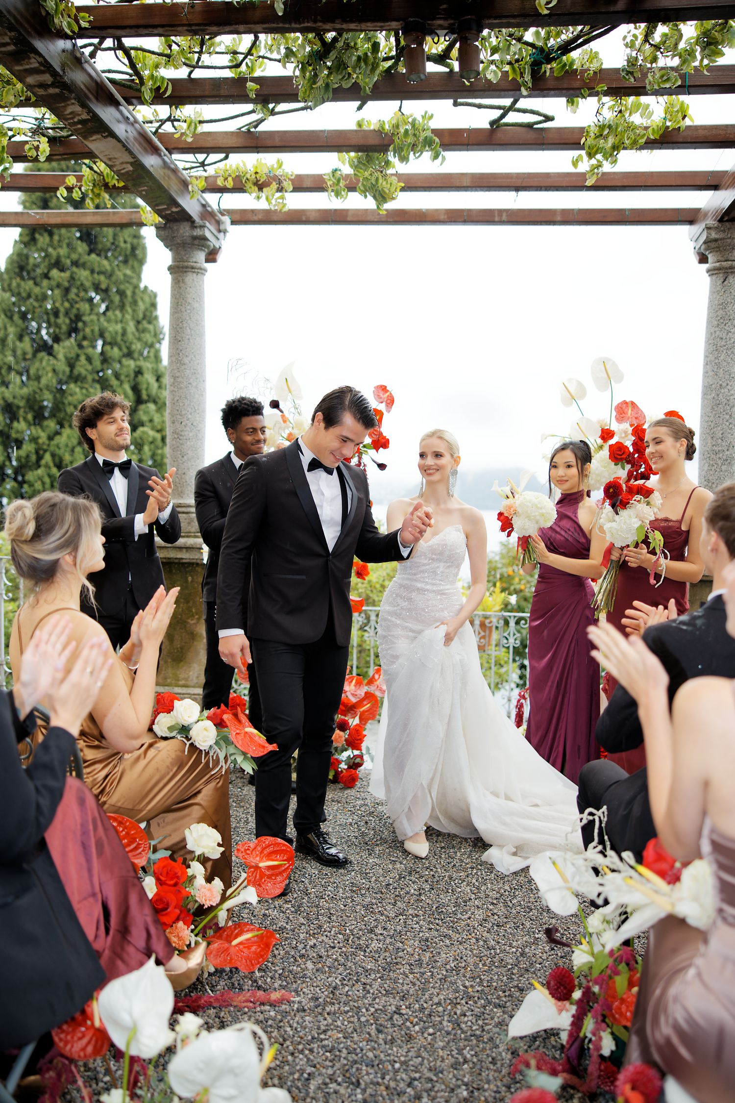 Wedding celebration under an elegant pergola decorated with vibrant red flowers and white accents.