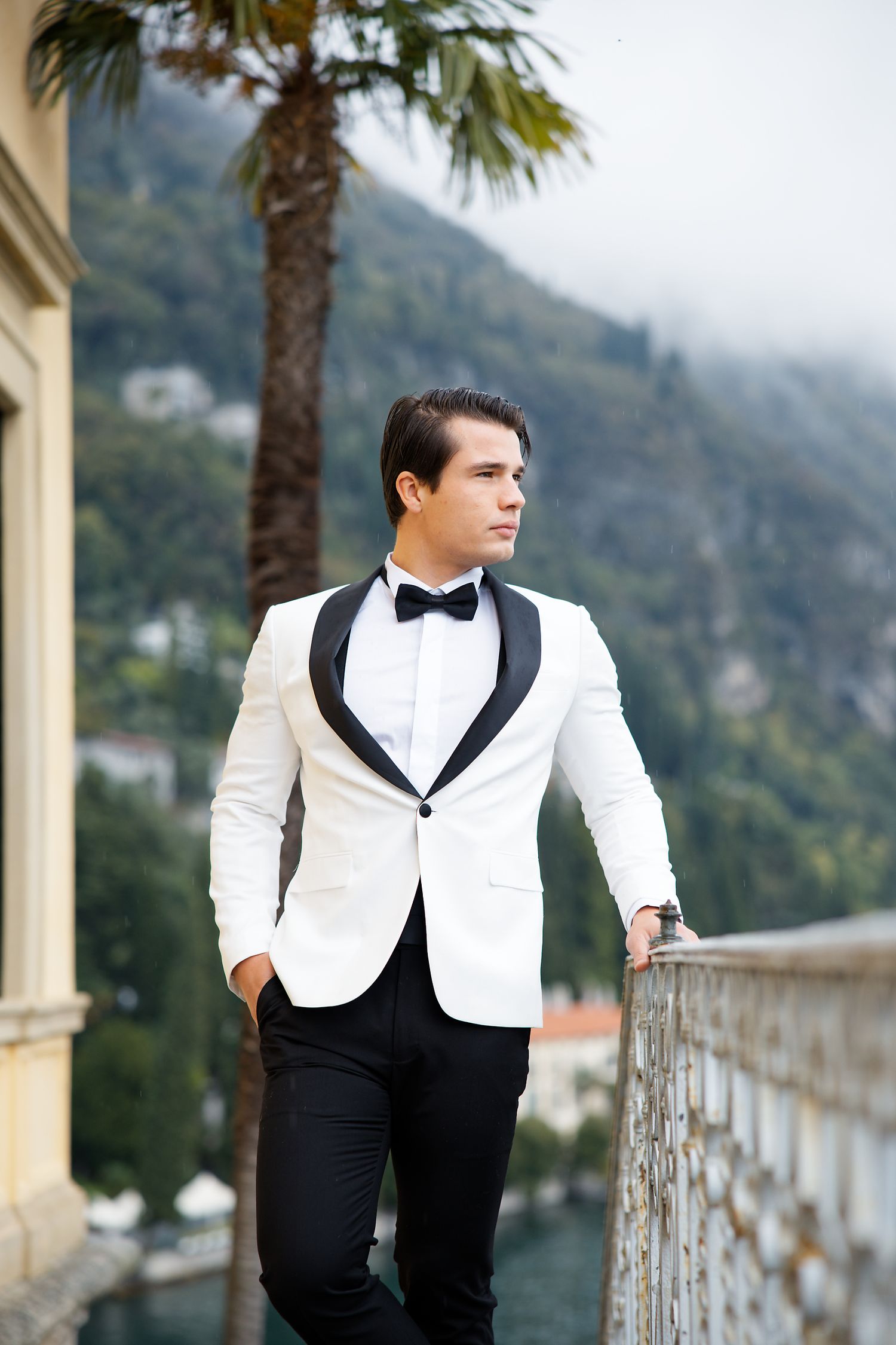 Elegant groom in white tuxedo poses on Mediterranean terrace with palm trees and mountain views.