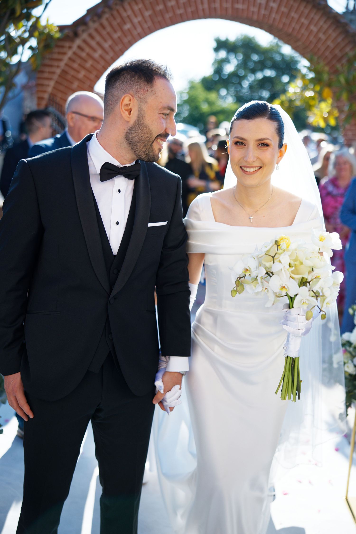 Newlyweds share joyful moment under brick archway with white orchid bouquet.