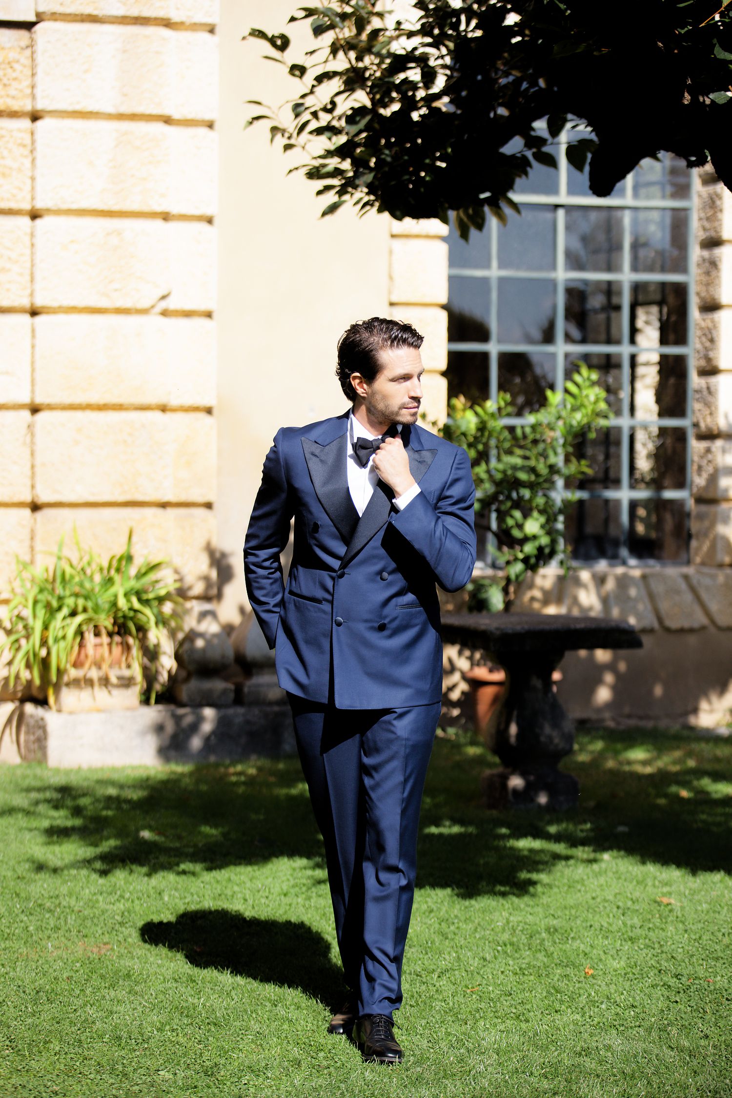 Groom in navy blue suit walking on grass with elegant architectural backdrop.