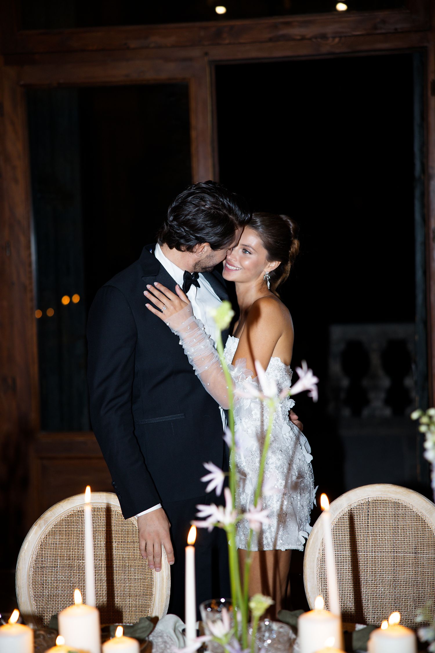 A romantic candlelit scene with a couple embracing at an elegant dinner table with floral arrangements.