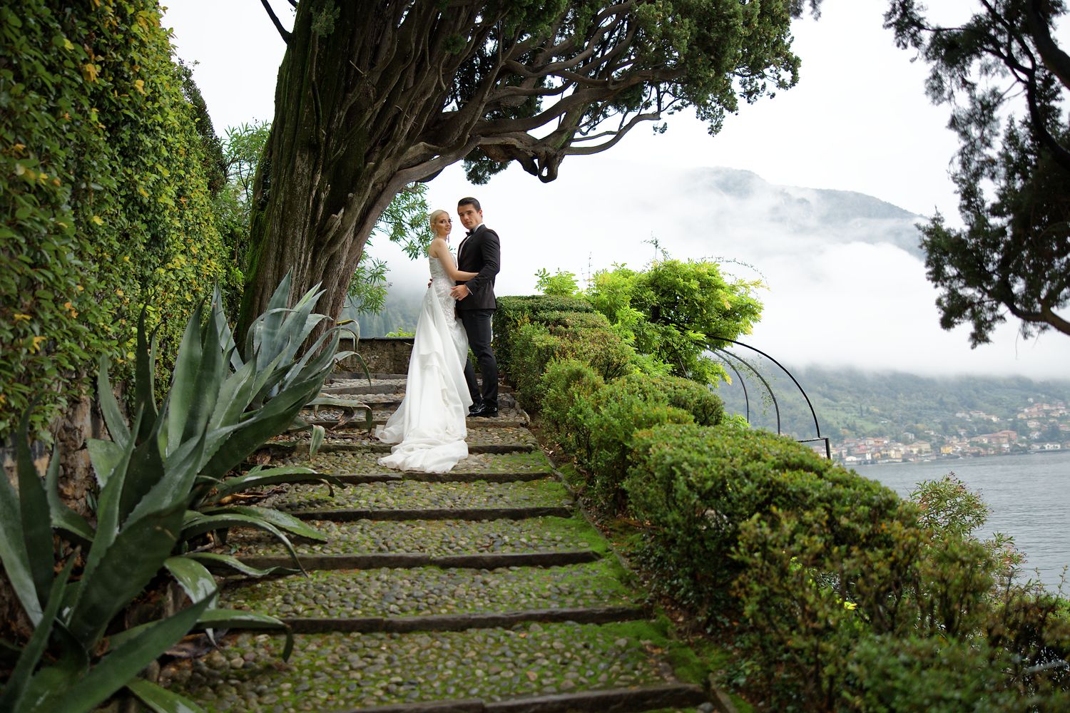 Newlyweds share a romantic moment on stone garden steps overlooking a lake vista.