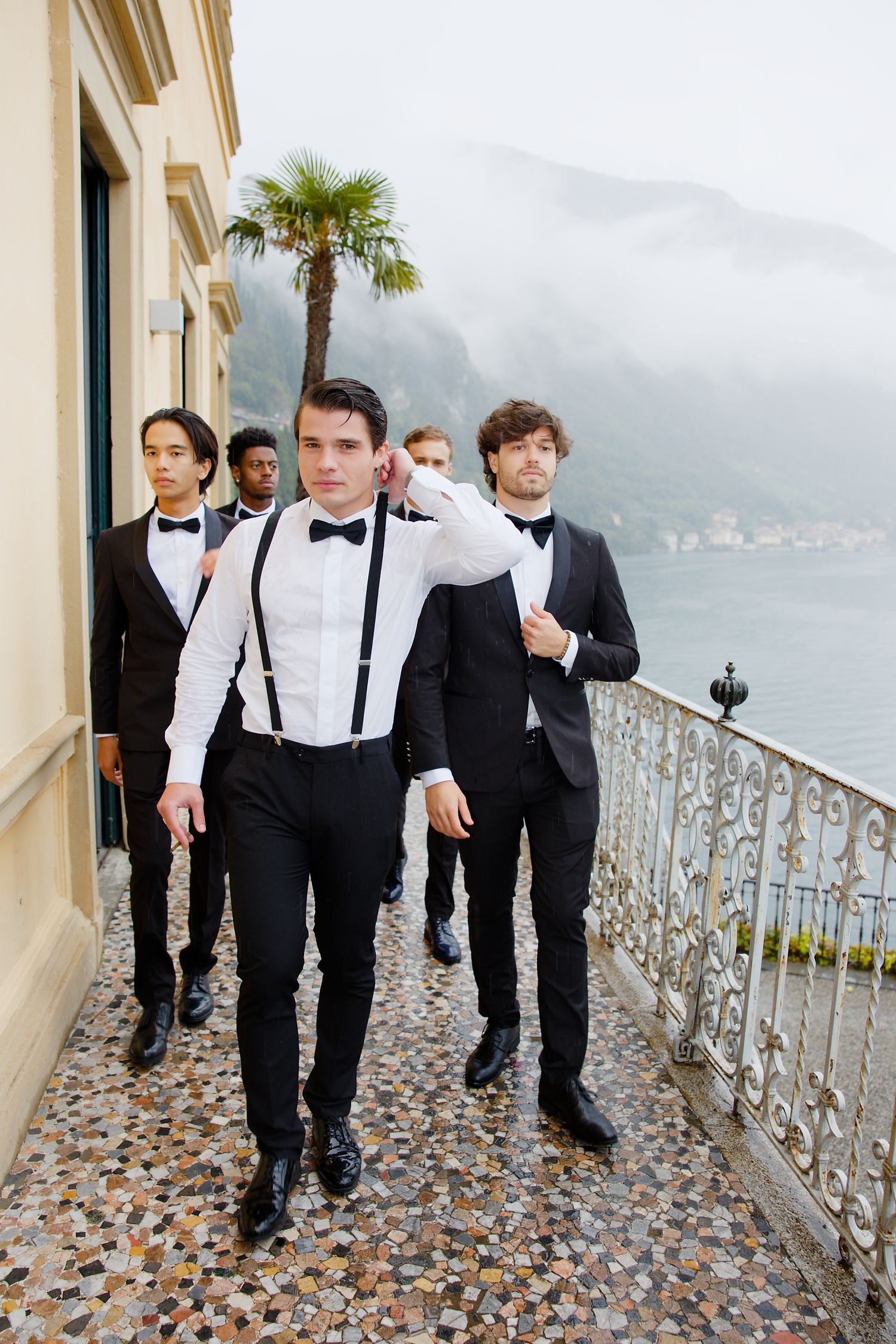 Stylish groomsmen in suspenders and bowties walking along Mediterranean balcony.