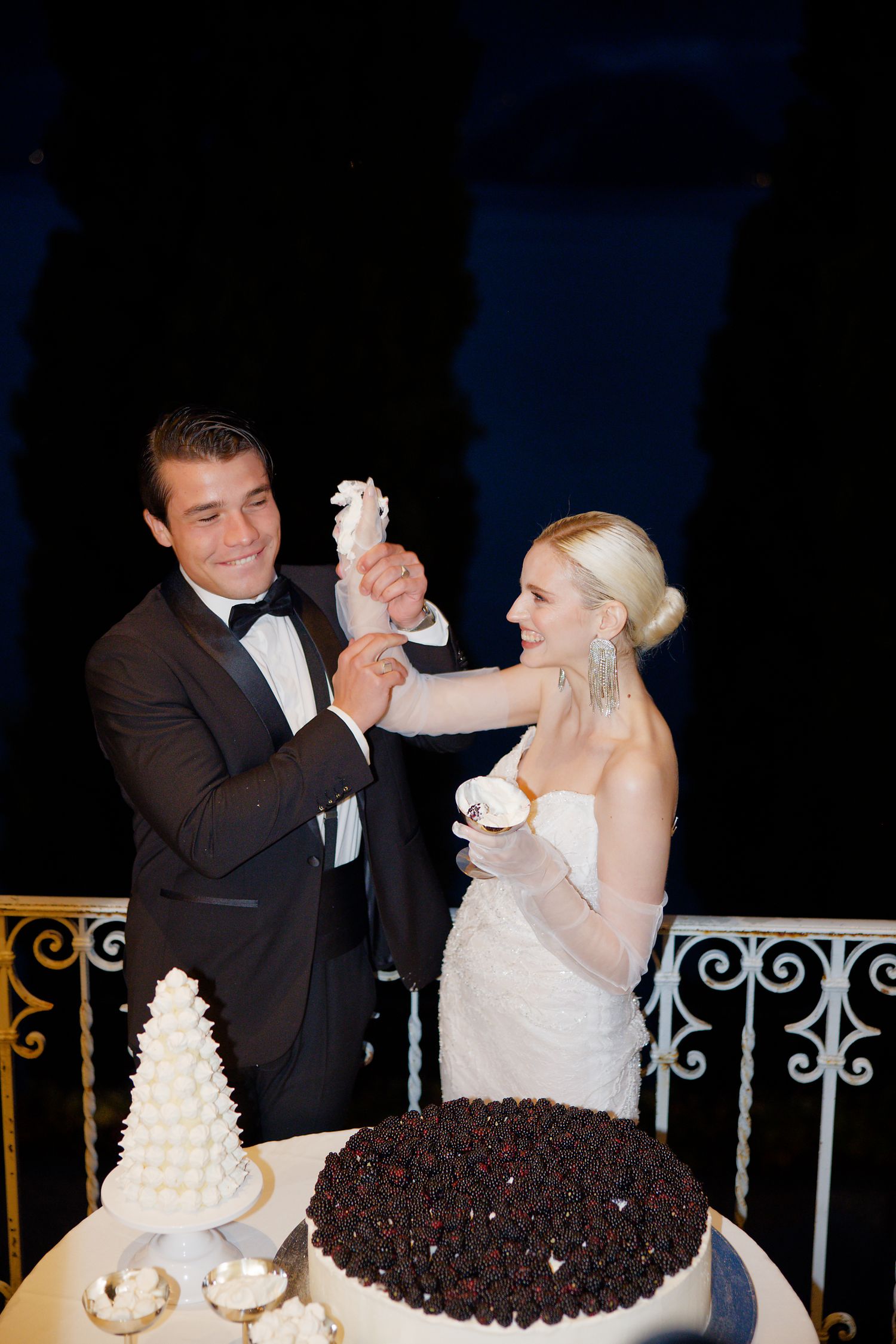 Wedding couple playfully feeds each other cake during their reception on a balcony at night.