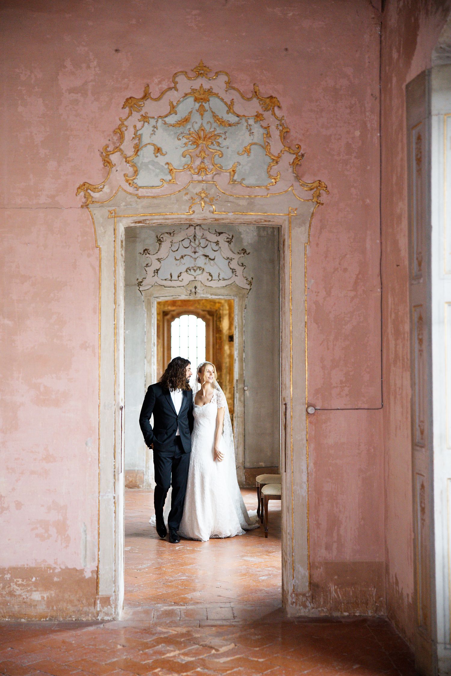 A bride and groom stand together in an ornate doorway framed by pink walls and baroque architectural details.