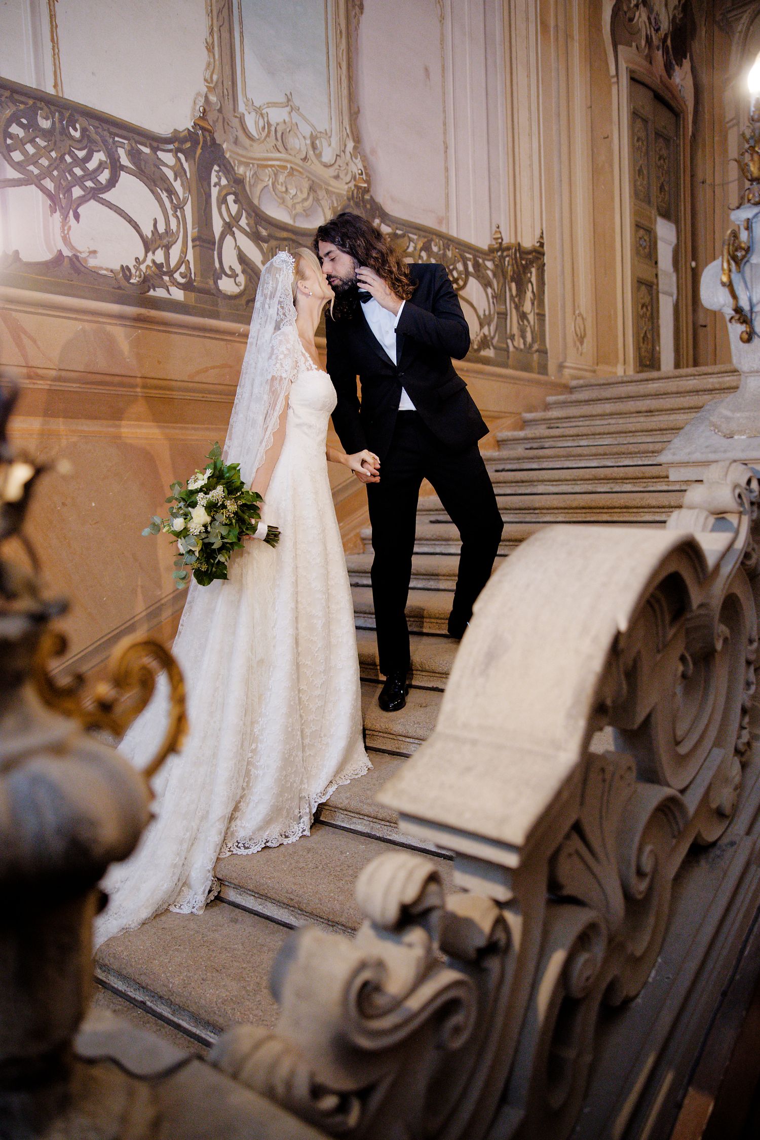 A romantic wedding couple poses on an ornate marble staircase in a luxurious palace setting.