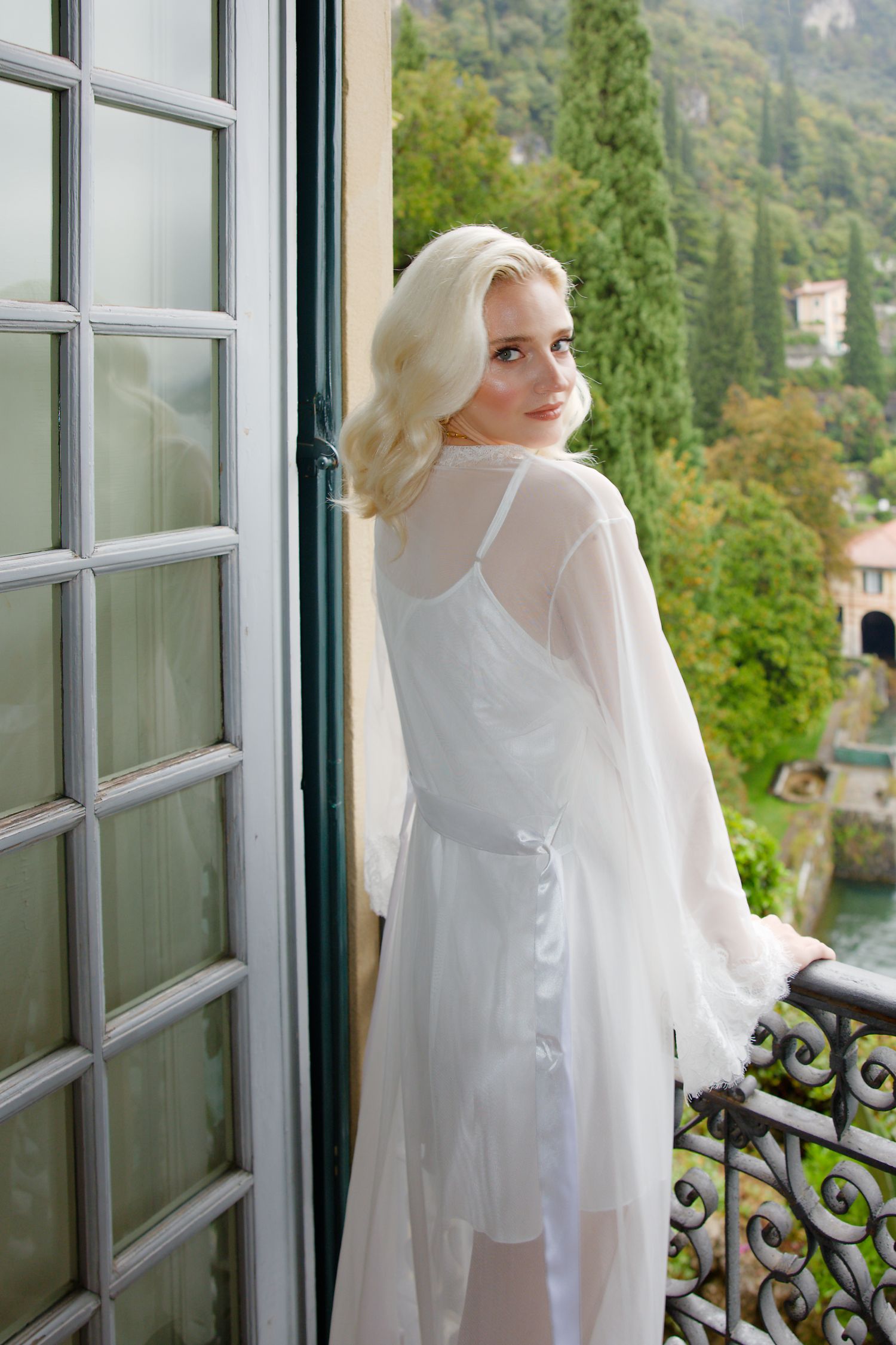 White flowy robe at a Mediterranean balcony overlooking scenic hillside villas and cypress trees.