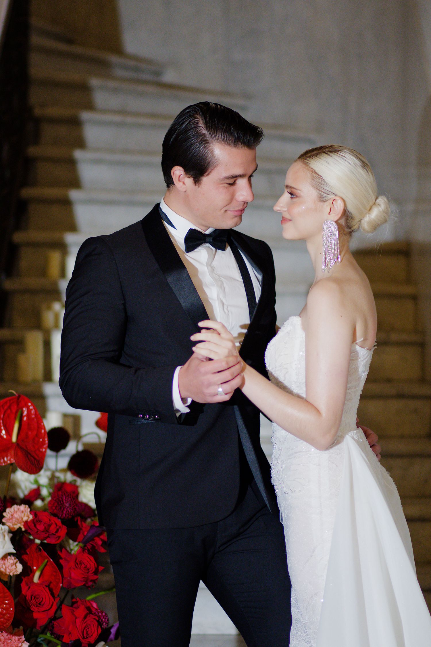 Romantic wedding moment as couple in formal attire dances near red floral arrangements.