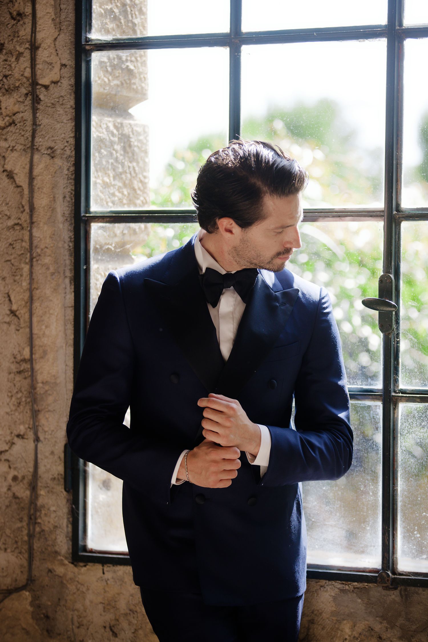 Groom adjusts his cufflink while standing by a window wearing a classic black tuxedo with bow tie.