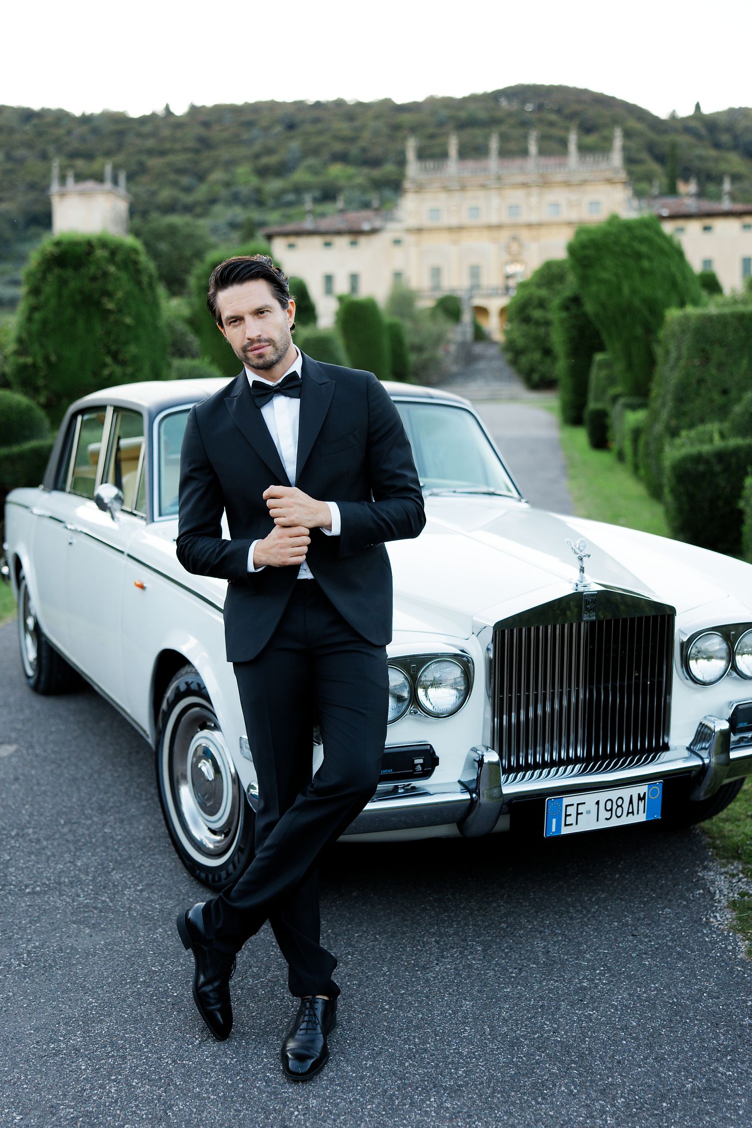 Elegant classic white Rolls-Royce parked in front of a grand Italian villa with manicured hedges in background.