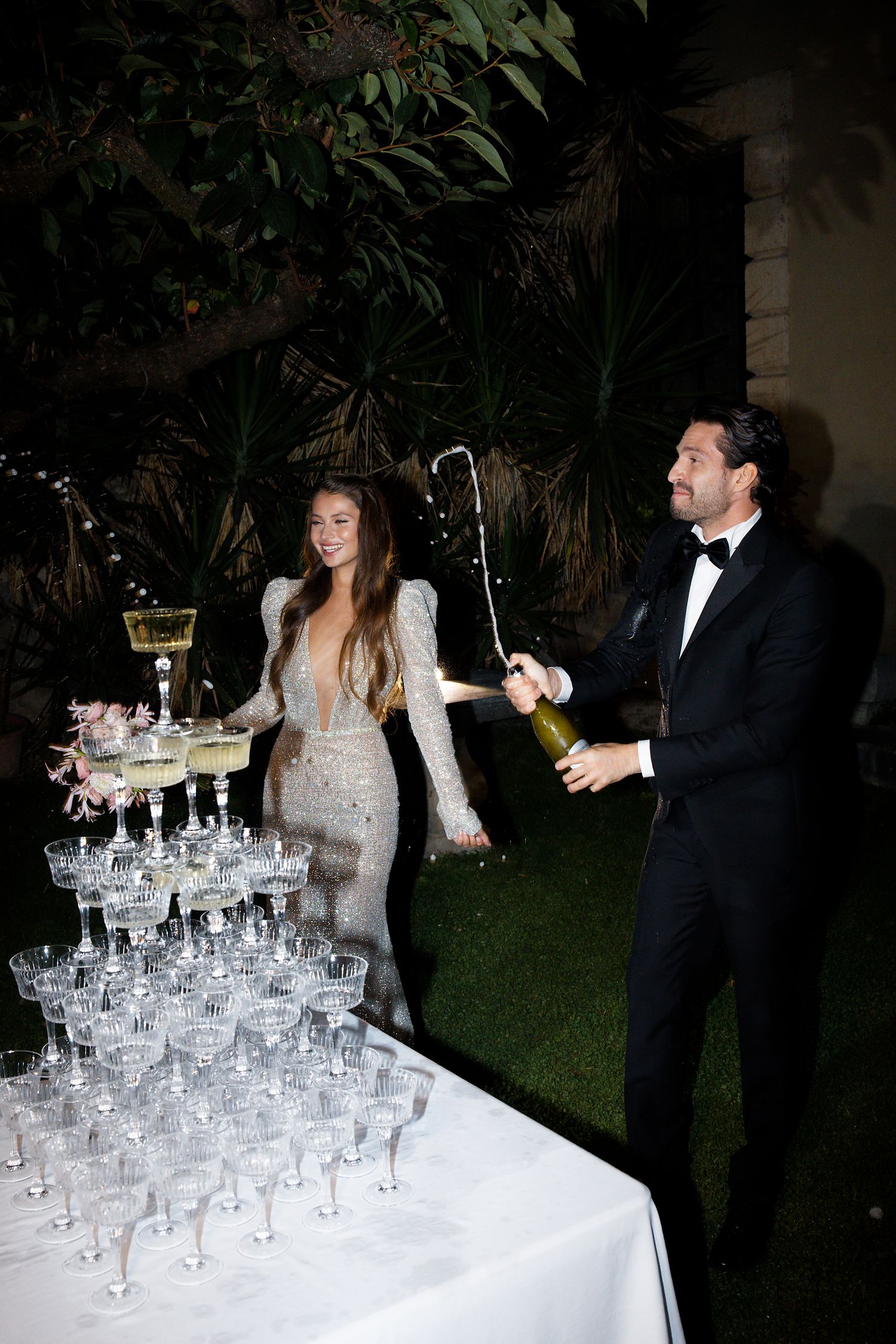 Couple pouring champagne into a tower of crystal glasses at an elegant outdoor wedding reception.