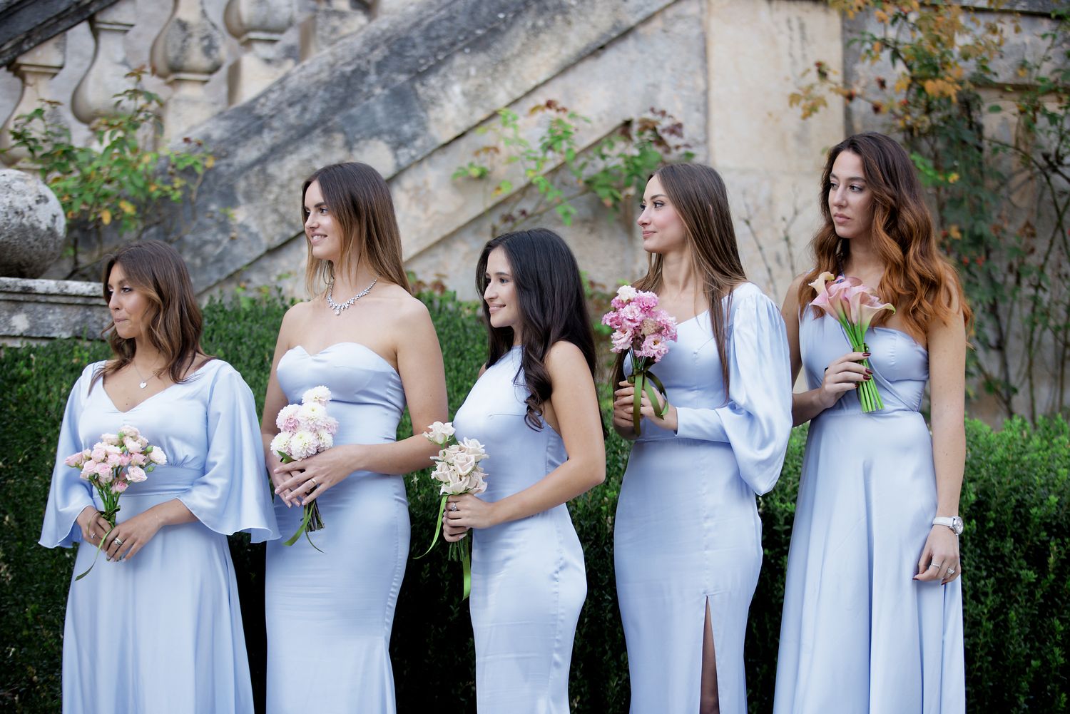 Five bridesmaids in light blue dresses holding pink bouquets stand together in a garden setting.
