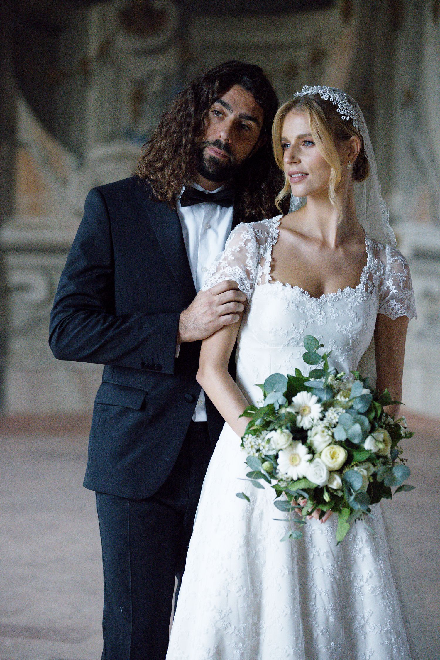 A wedding portrait of a couple in formal attire standing against an elegant palace interior.