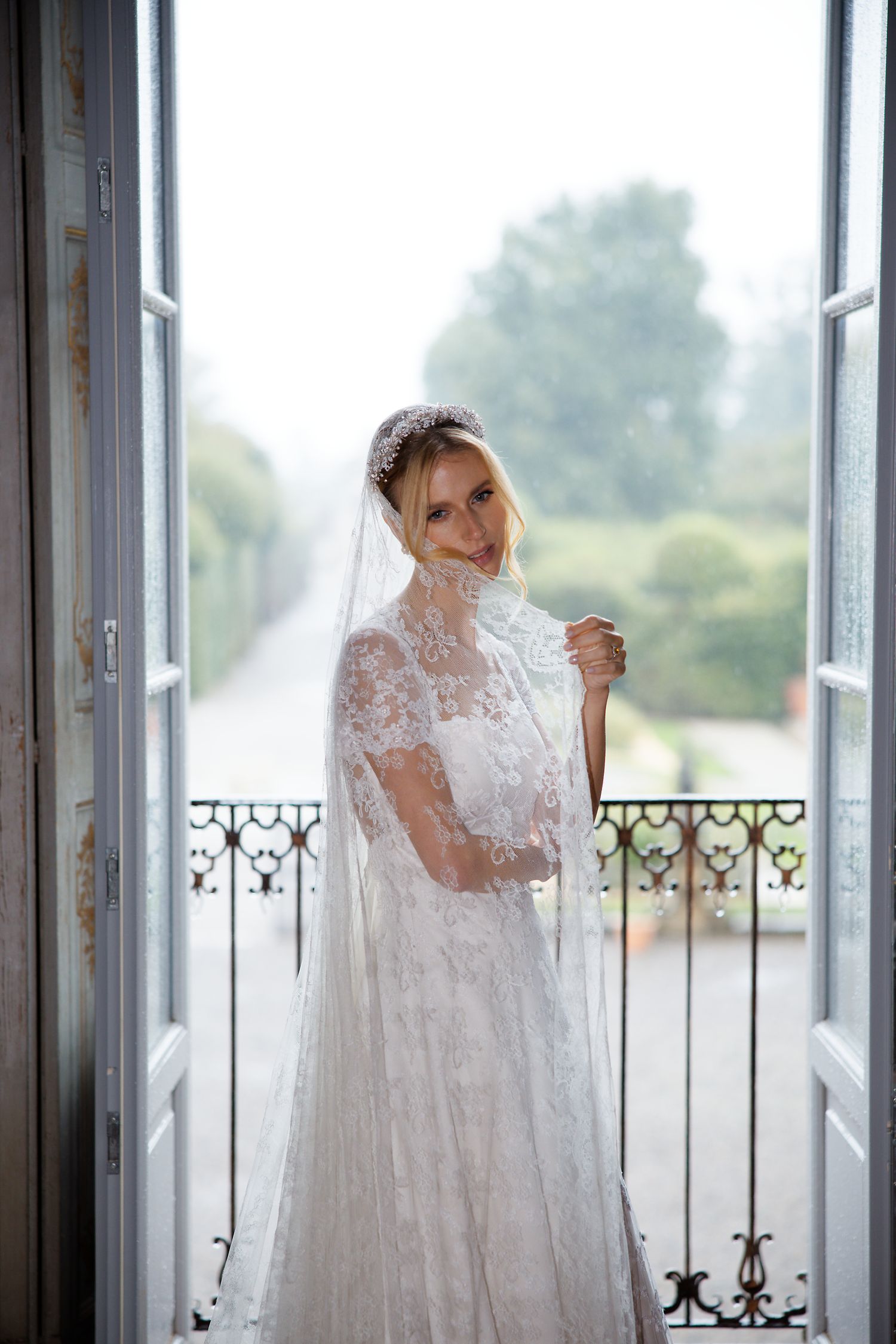 Ethereal bridal portrait with lace gown and veil framed by French doors opening to a balcony.