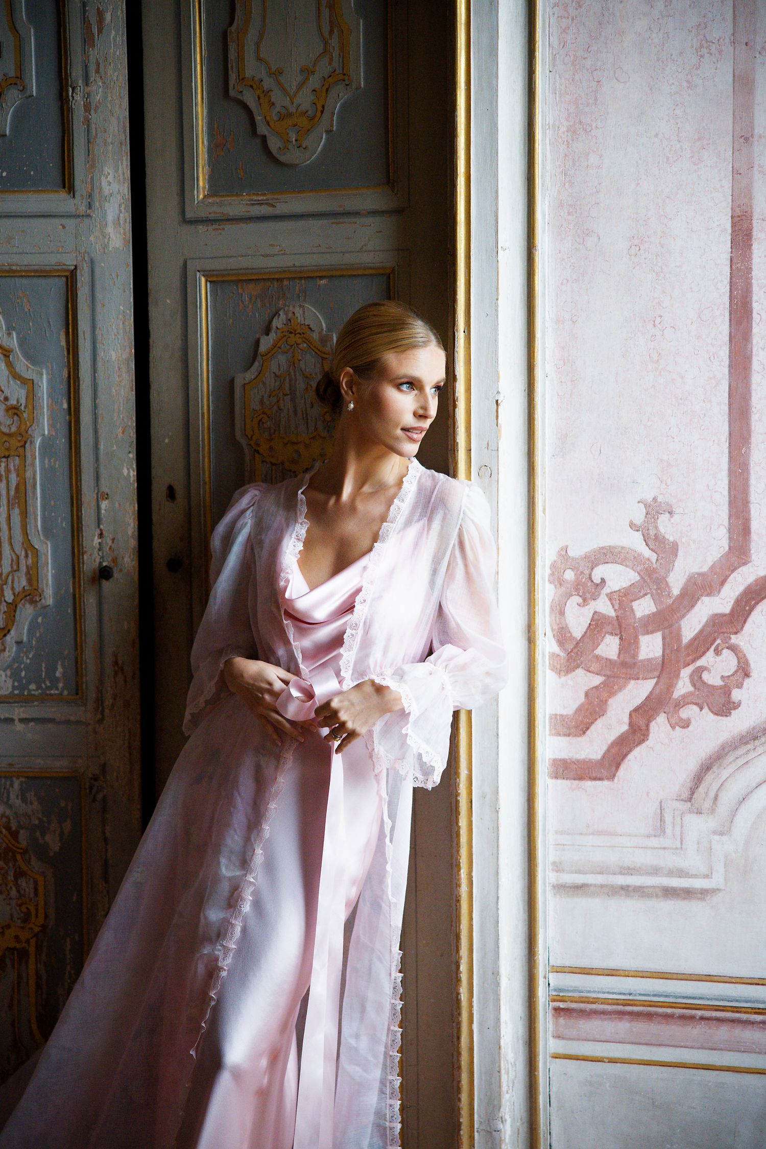 An elegant figure in a pink silk robe poses by an ornate vintage door with gilded details.