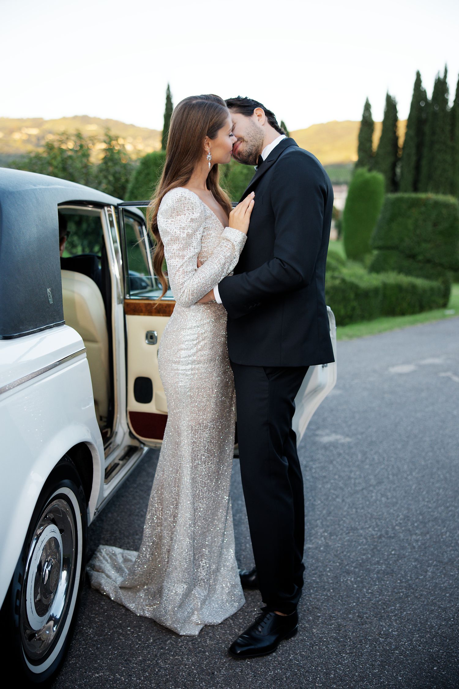 Newlywed couple sharing an intimate moment beside a vintage car on their wedding day.