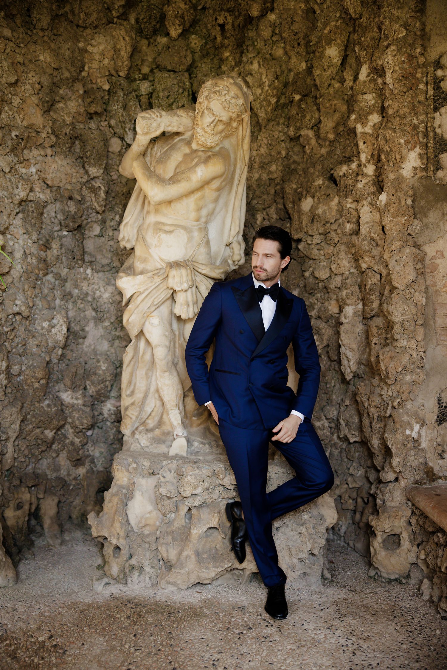 A groom in a navy blue tuxedo poses against a stone wall with a classical statue in an elegant wedding photo.