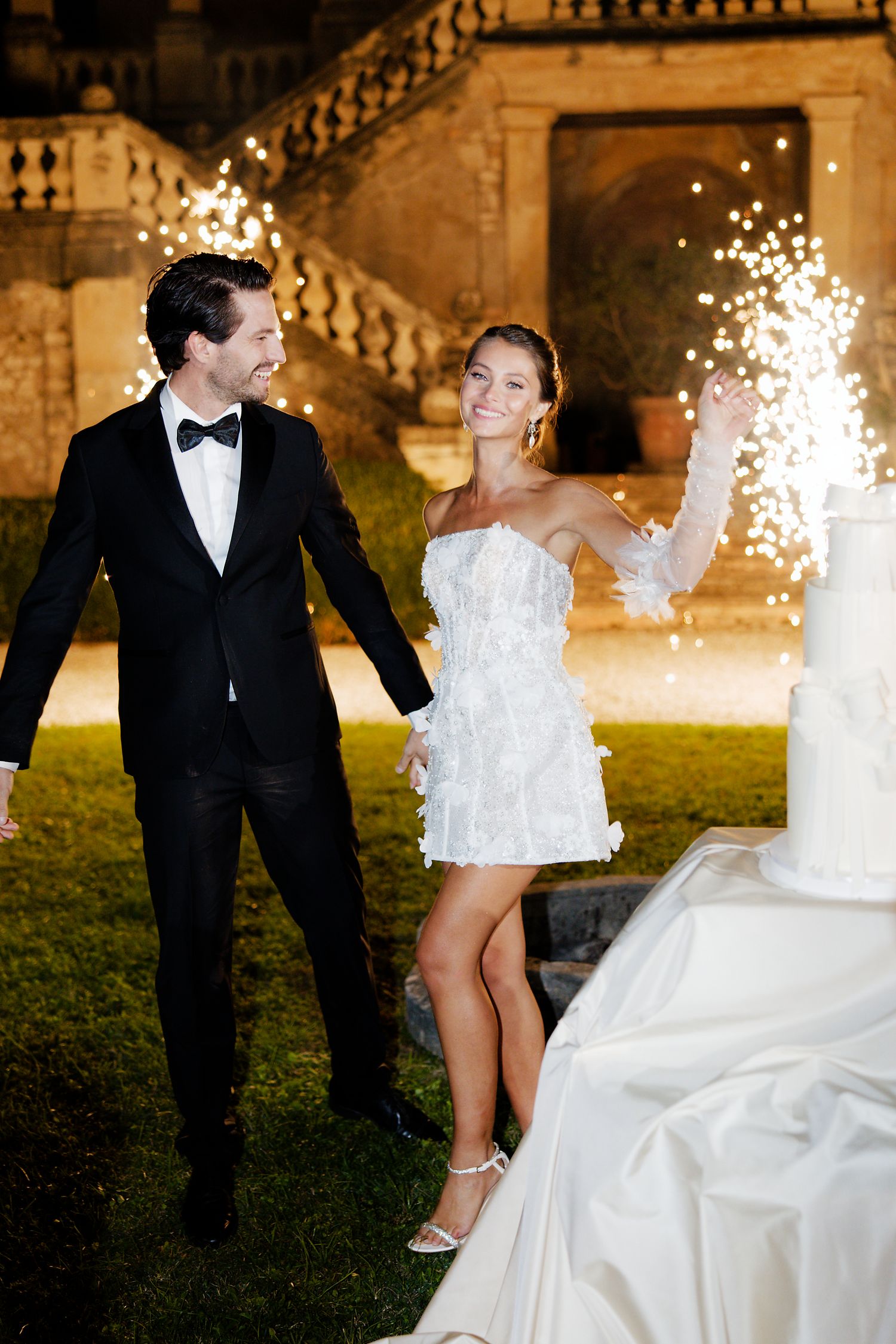 Couple celebrating with sparklers next to their white wedding cake in garden setting.